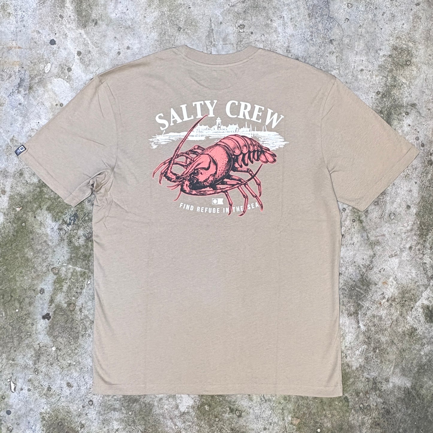 A beige short-sleeve t-shirt laid flat on a textured concrete surface, featuring a large graphic print on the back with the words 'SALTY CREW' in white and a red lobster illustration.