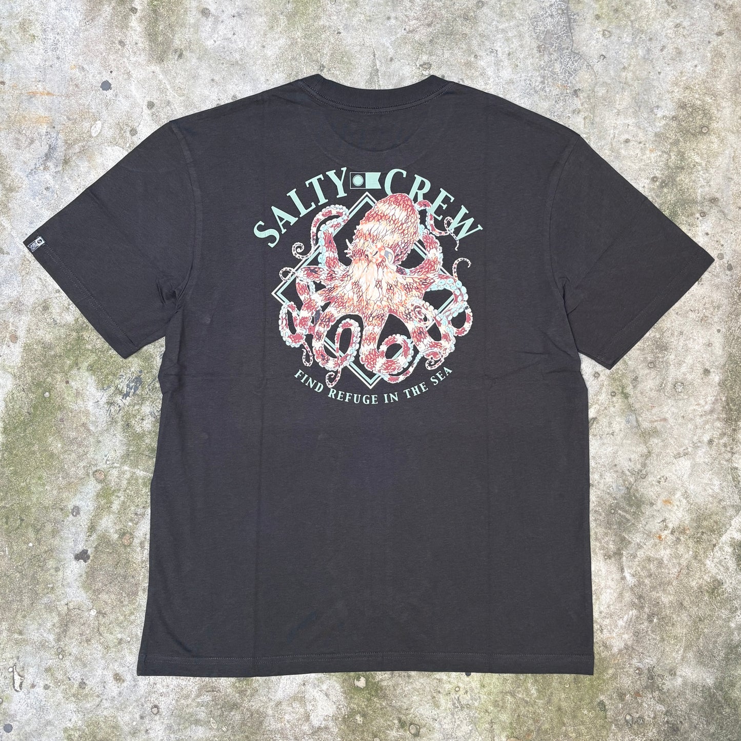 A black short-sleeve t-shirt laid flat on a textured concrete surface, featuring a large back graphic with an octopus, anchor, and the words 'SALTY CREW' and 'FIND REFUGE IN THE SHORE'.