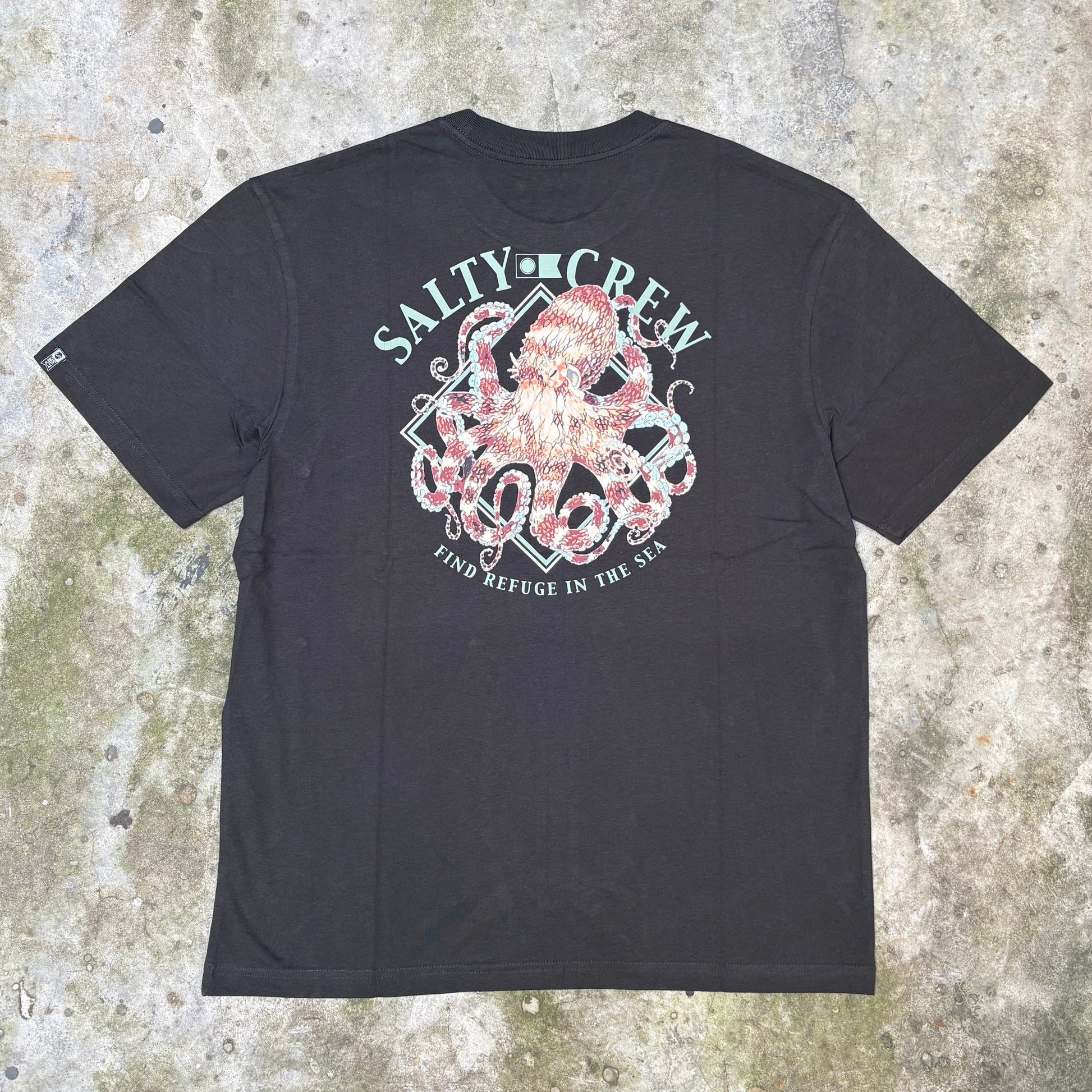 A black short-sleeve t-shirt laid flat on a textured concrete surface, featuring a large back graphic with an octopus, anchor, and the words 'SALTY CREW' and 'FIND REFUGE IN THE SHORE'.