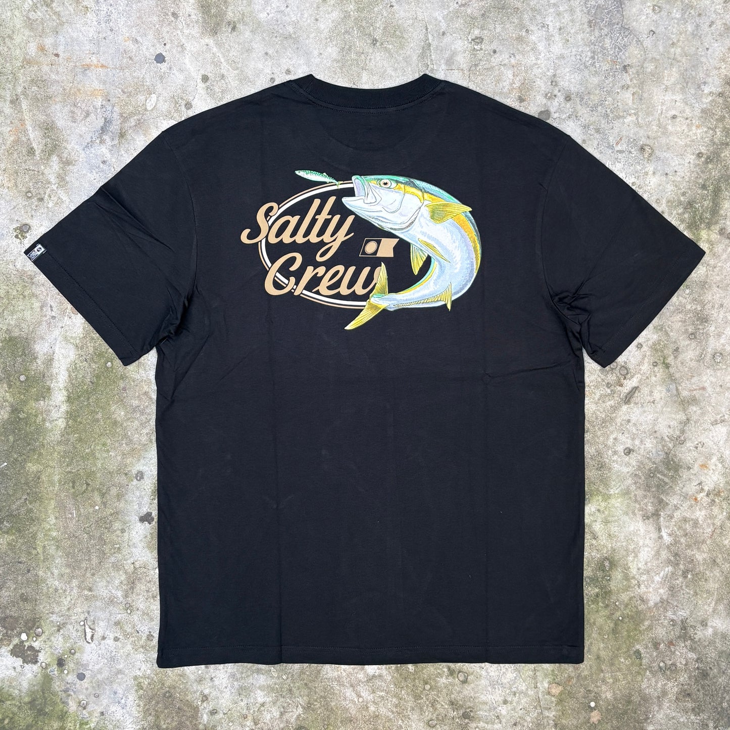 A black short-sleeve t-shirt laid flat on a textured concrete surface, featuring a large graphic on the chest of a yellow and blue mahi-mahi fish with the text 'Salty Crew' in gold and white.