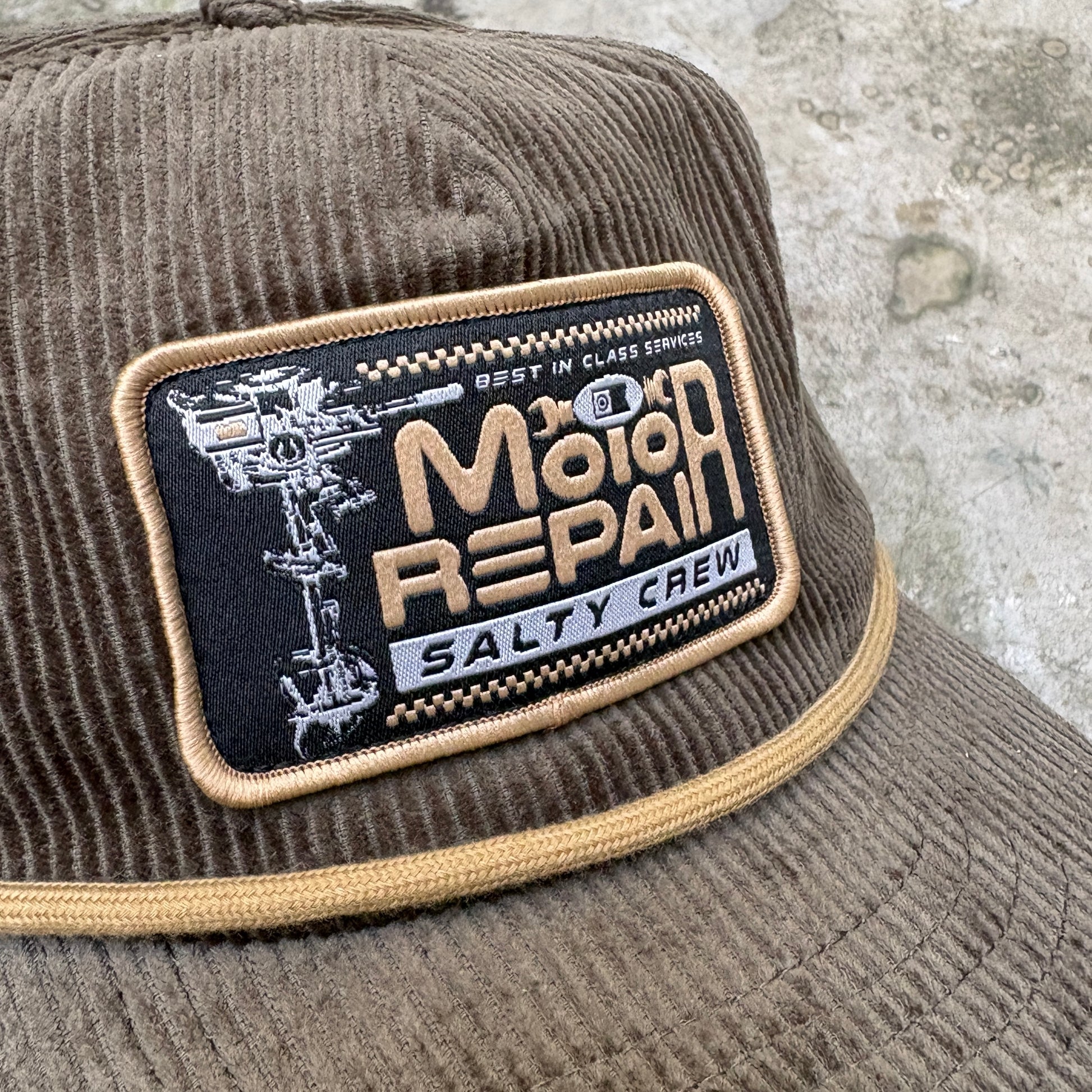 A close-up of a brown corduroy trucker hat featuring a rectangular embroidered patch on the front. The patch reads 'Moto Repair' and 'Salty Crew' with a black background and tan lettering, bordered in tan corduroy trim.