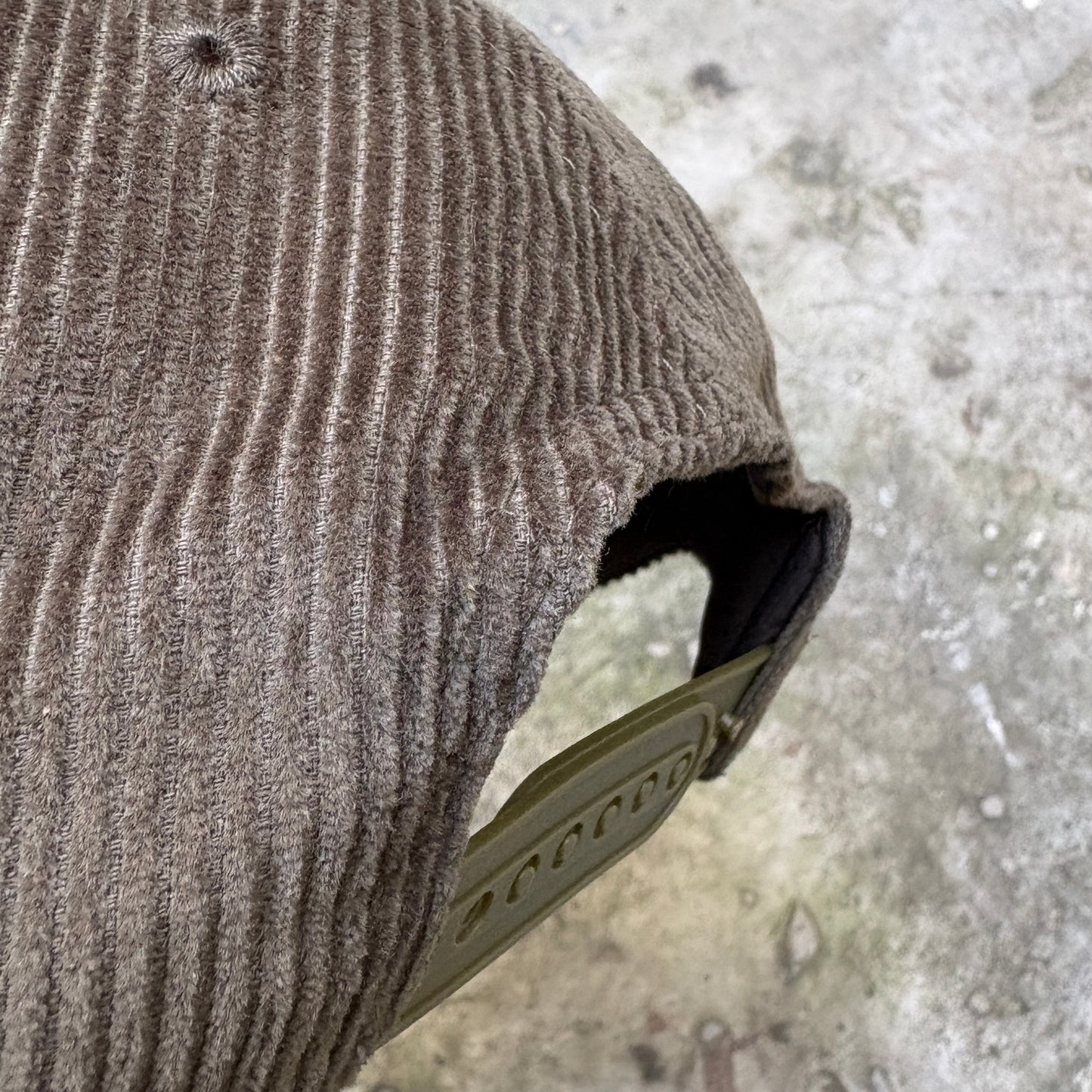 Close-up of a brown corduroy cap showing the ribbed fabric texture and a greenish olive-colored plastic visor edge against a concrete ground background.