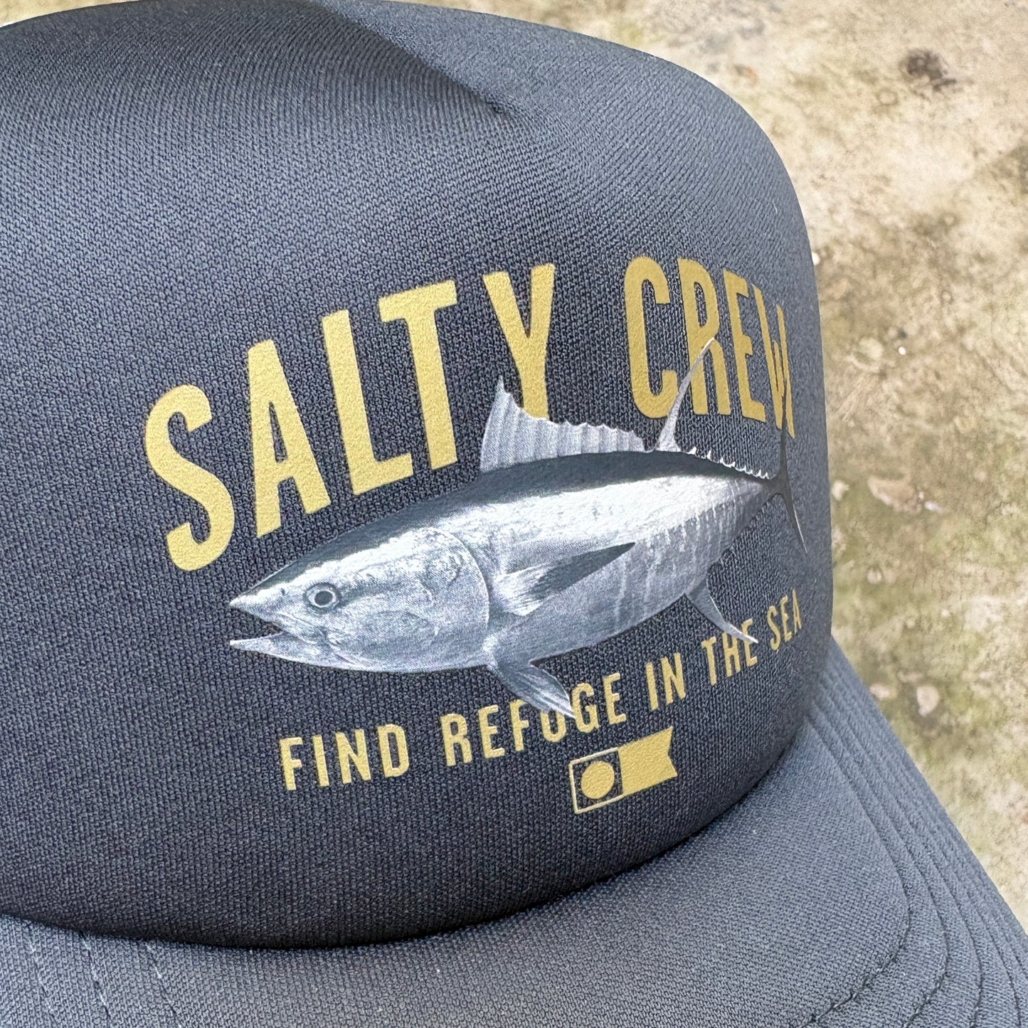 A dark gray trucker-style cap featuring a large graphic print of a tuna fish and the text 'SALTY CREW' and 'FIND REFUGE IN THE SEA' in yellow, photographed outdoors against a sandy ground.