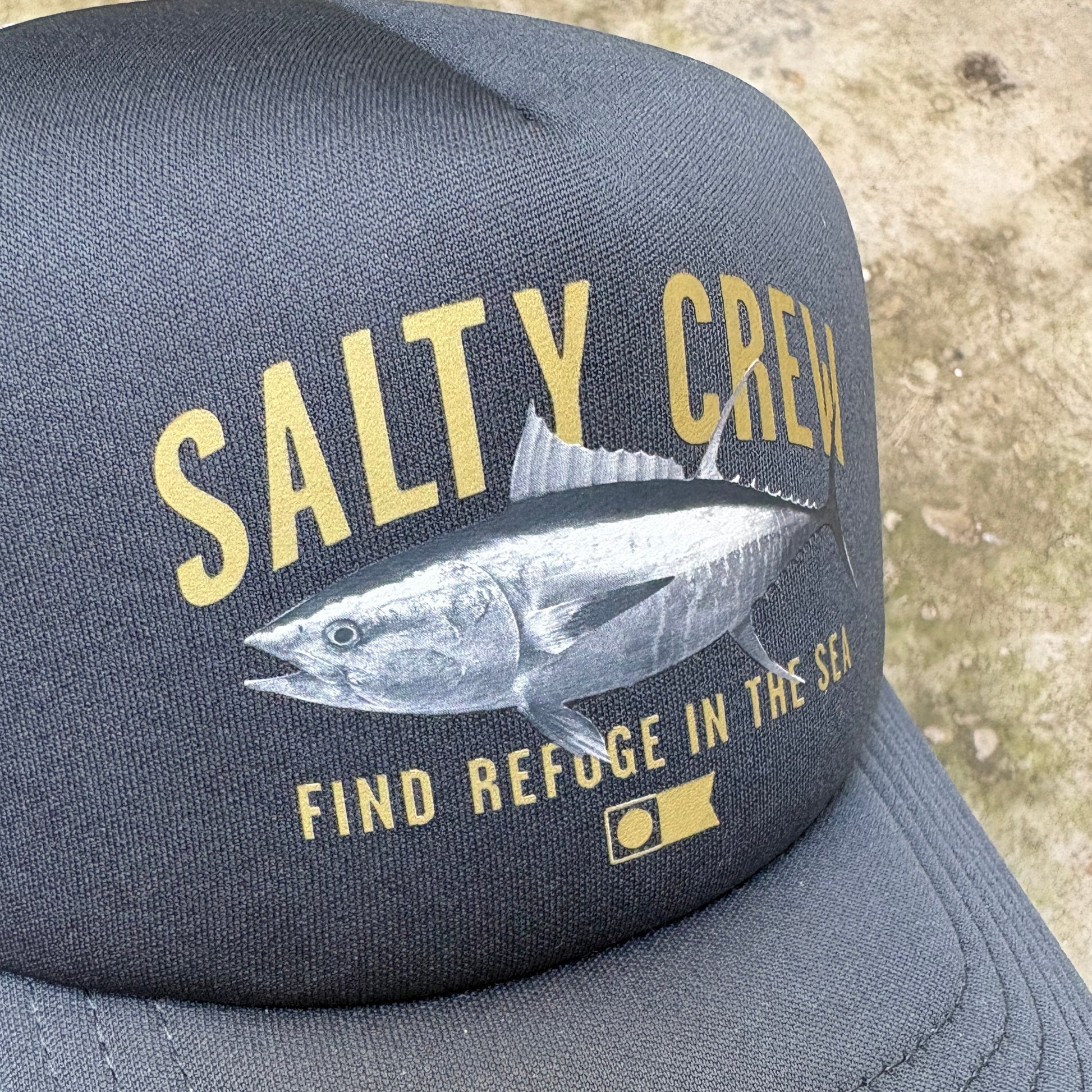 A dark gray trucker-style cap featuring a large graphic print of a tuna fish and the text 'SALTY CREW' and 'FIND REFUGE IN THE SEA' in yellow, photographed outdoors against a sandy ground.