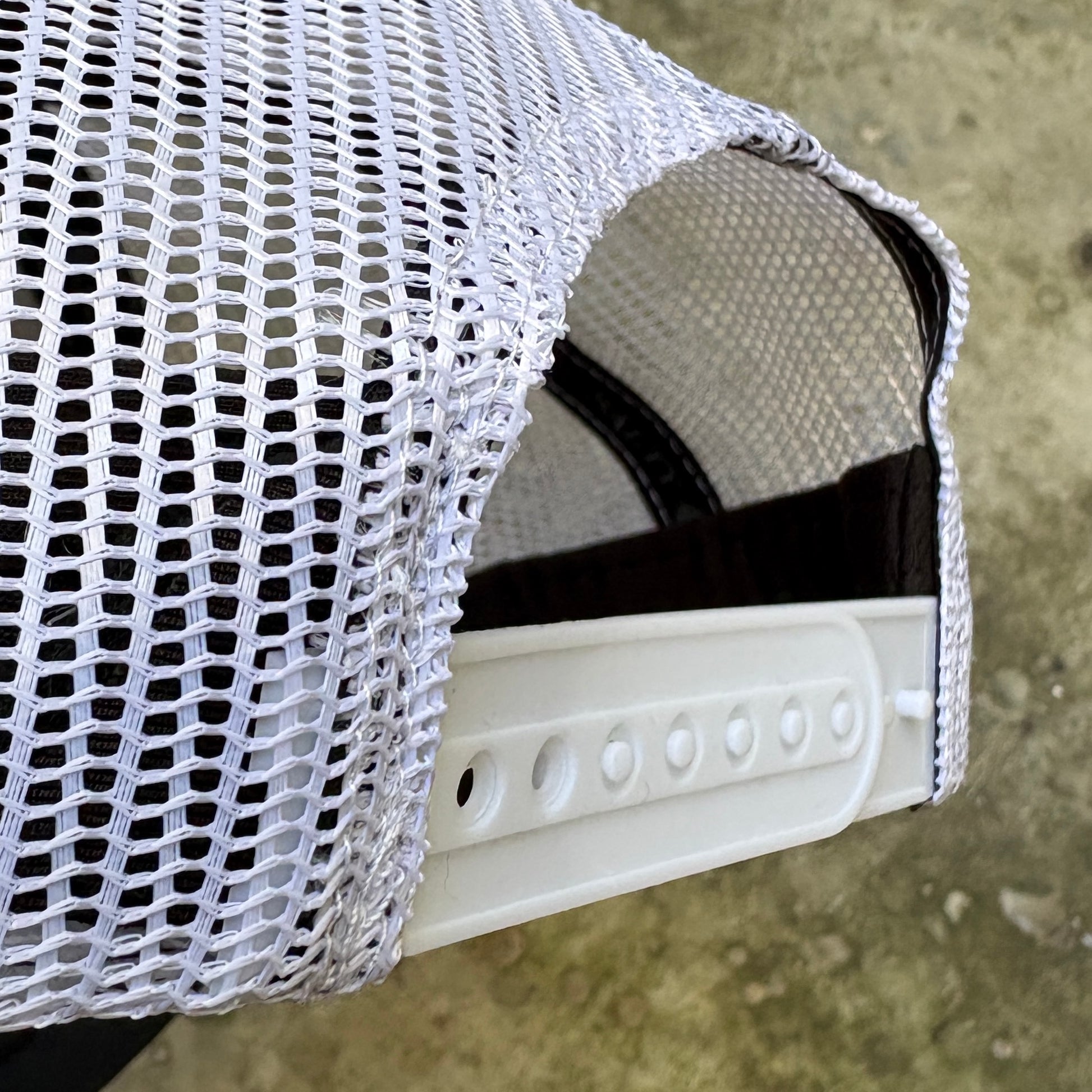 Close-up of a white mesh trucker hat showing the side and back panel with adjustable ventilation holes and a white plastic visor.