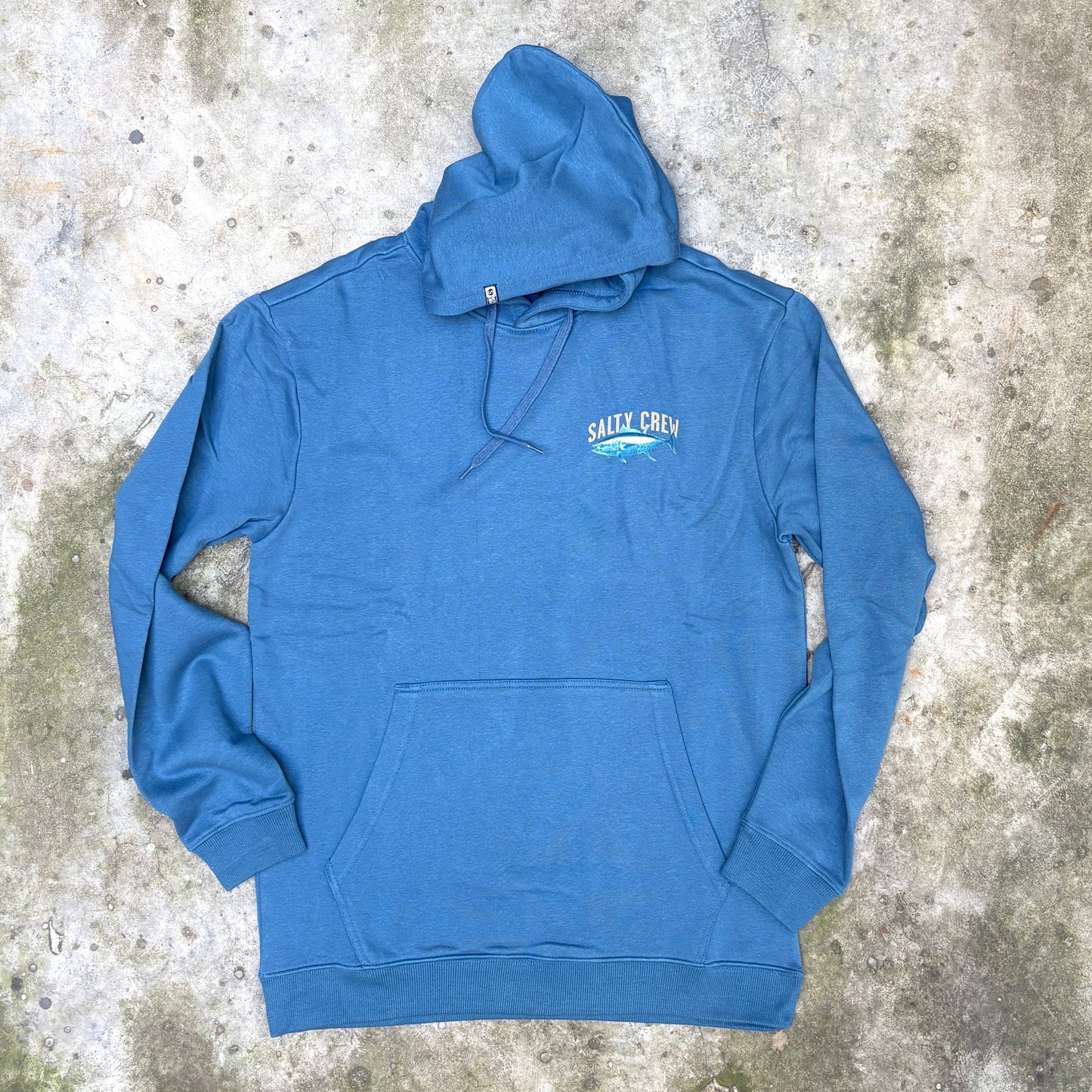 A blue pullover hoodie with a small 'SHARK CREW' graphic on the left chest, laid flat on a textured concrete surface.
