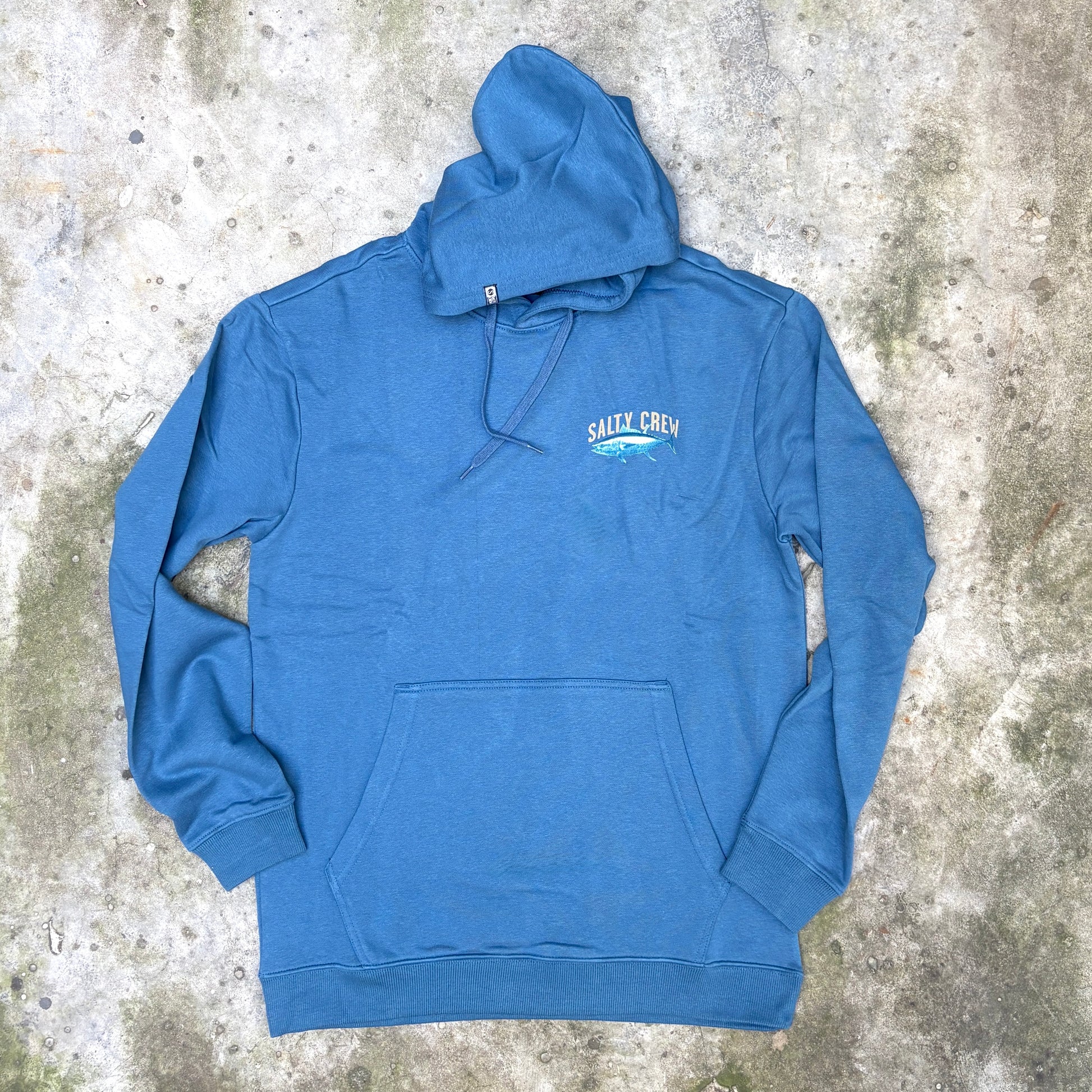 A blue pullover hoodie with a small 'SHARK CREW' graphic on the left chest, laid flat on a textured concrete surface.