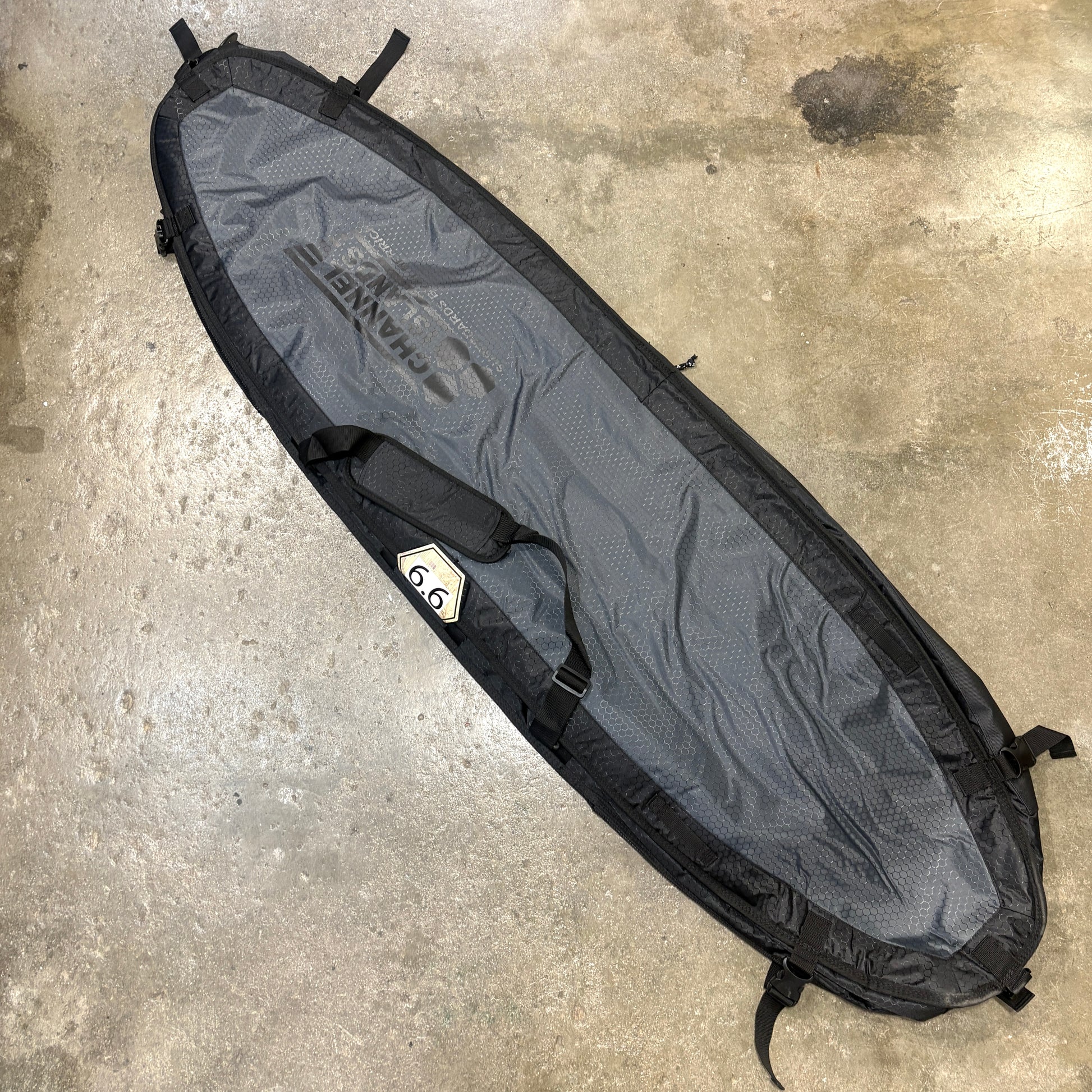 A long gray and black surfboard carrying bag laid diagonally on a concrete floor. The bag has black straps, buckles, and trim, and a visible printed brand name that appears to read 'HARNESS'.