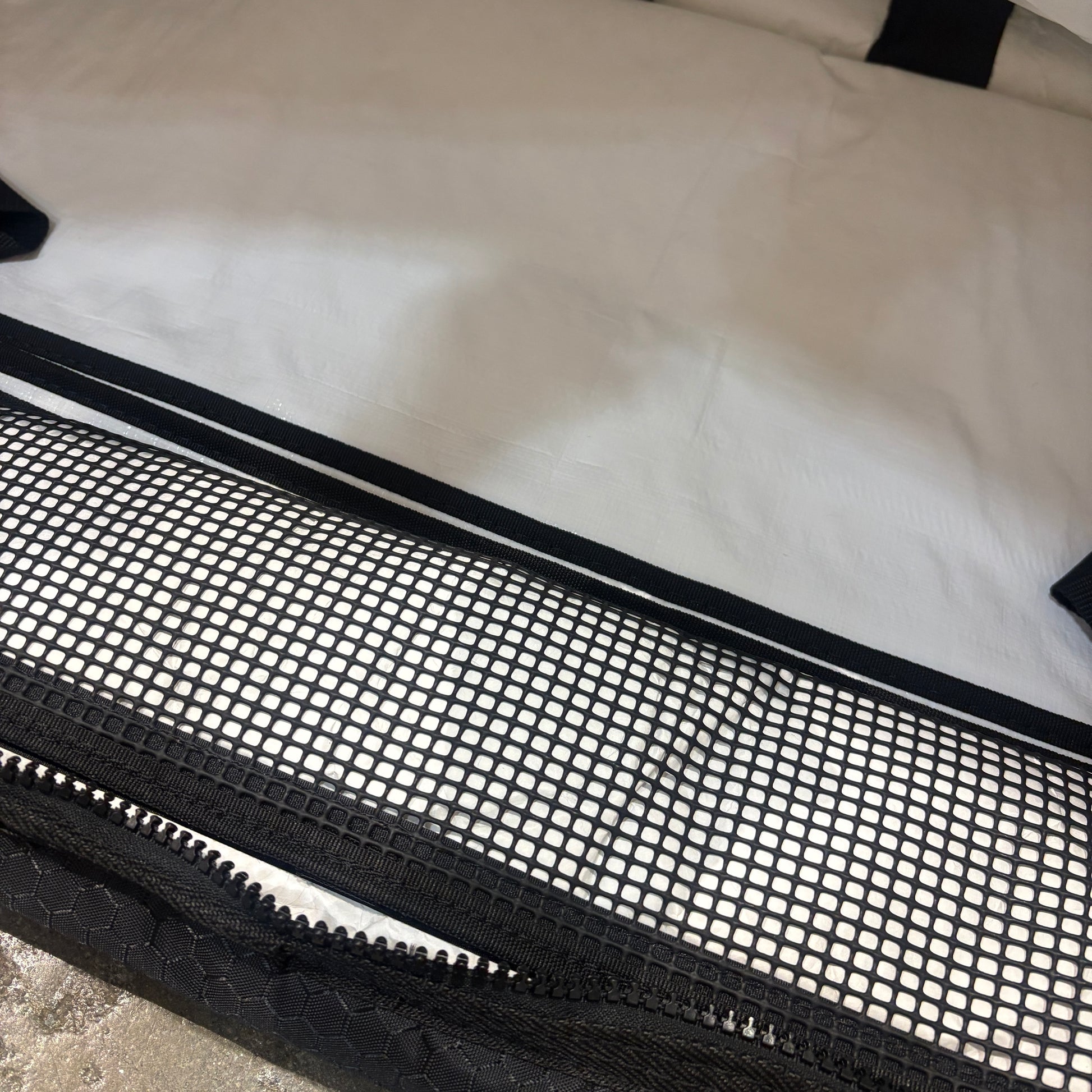 Close-up of a black mesh zippered storage bag or luggage sleeve laid over a white suitcase or bag, showing the black zipper, mesh pattern, and trim.