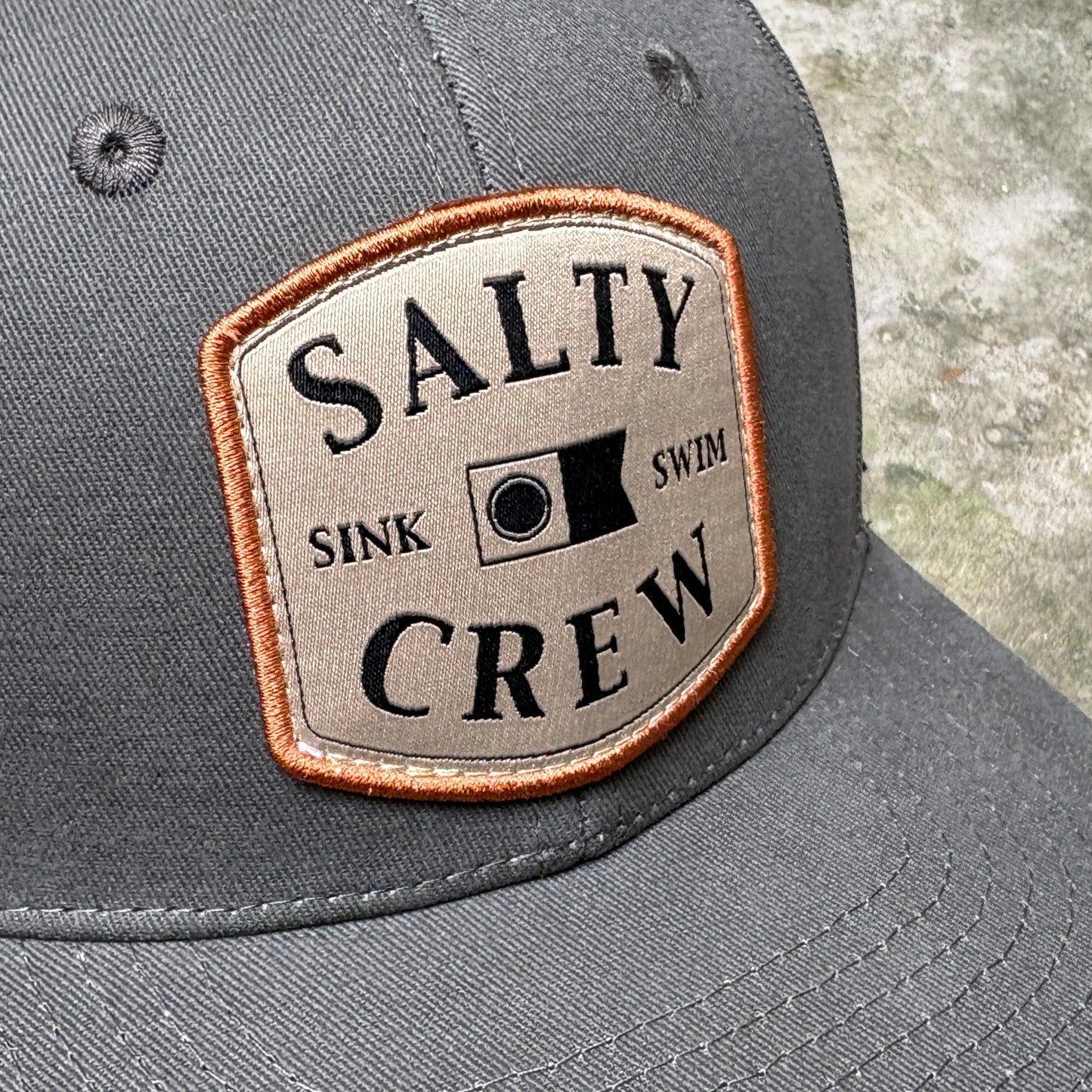 Close-up of a dark gray baseball cap featuring an embroidered patch on the front. The patch is beige with a copper-orange stitched border and black text reading 'SALTY CREW' with 'SINK SWIM' and a flag graphic in the center.