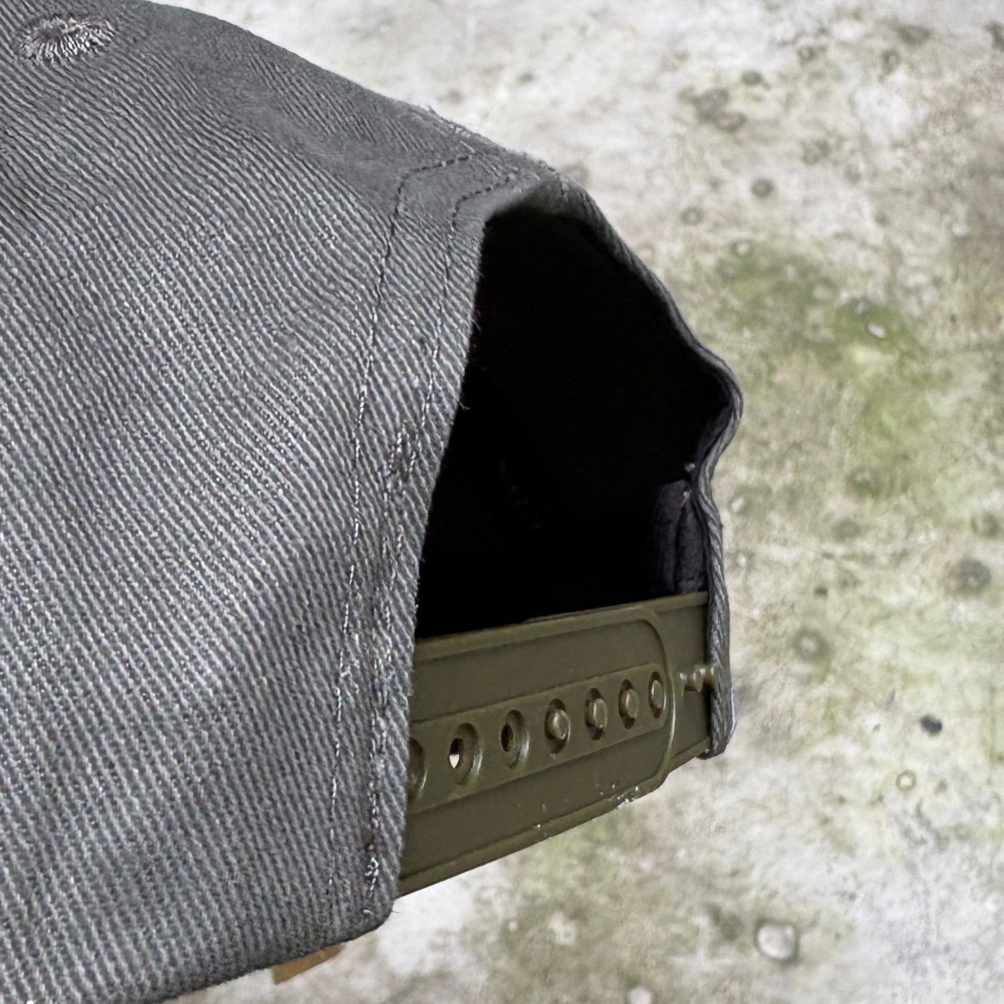 Close-up of a dark gray canvas pouch or sleeve with a stitched opening, showing a dark interior and a metal clip or bracket attached along the edge, set against a mottled concrete surface.