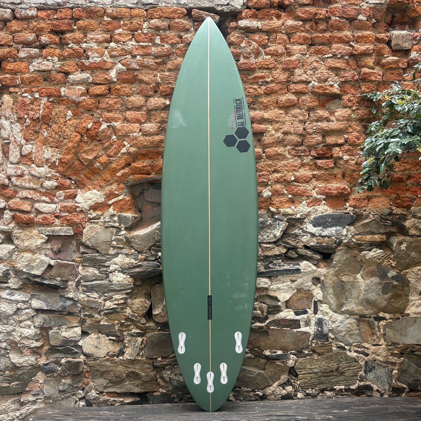 A green surfboard with black graphics, standing vertically against a rustic brick and stone wall.