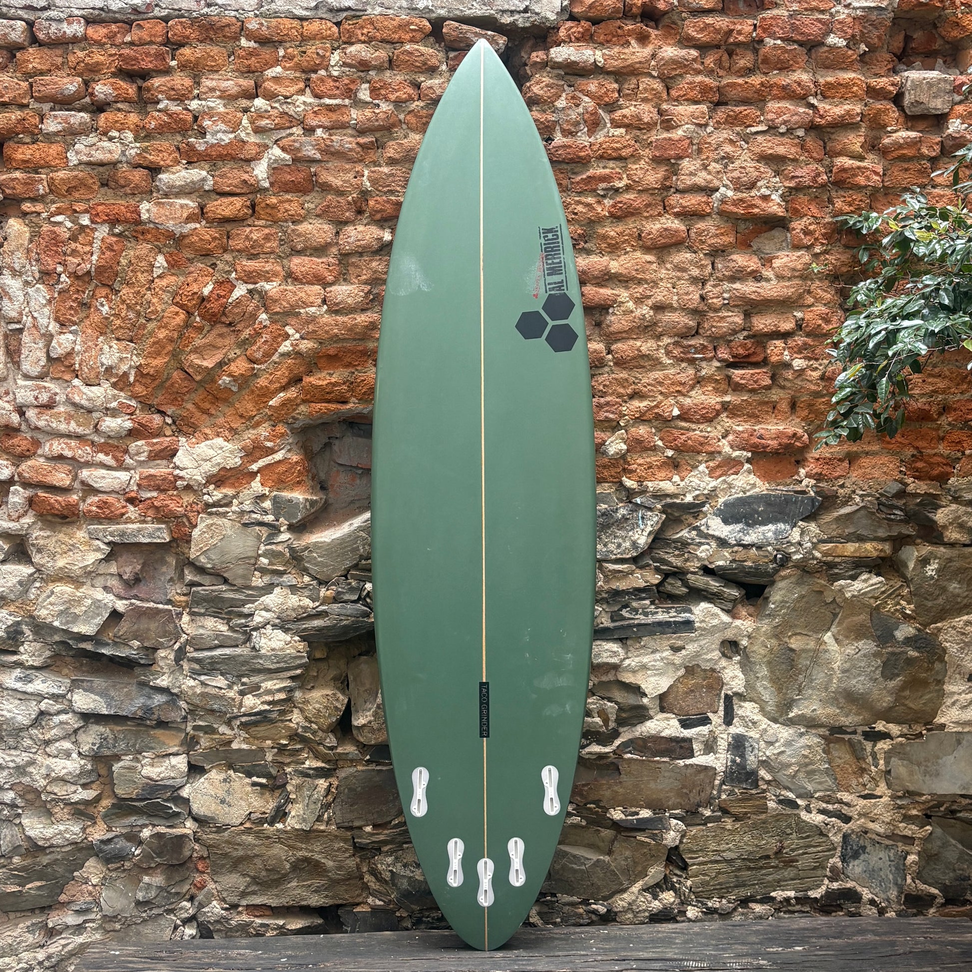 A green surfboard with black graphics, standing vertically against a rustic brick and stone wall.