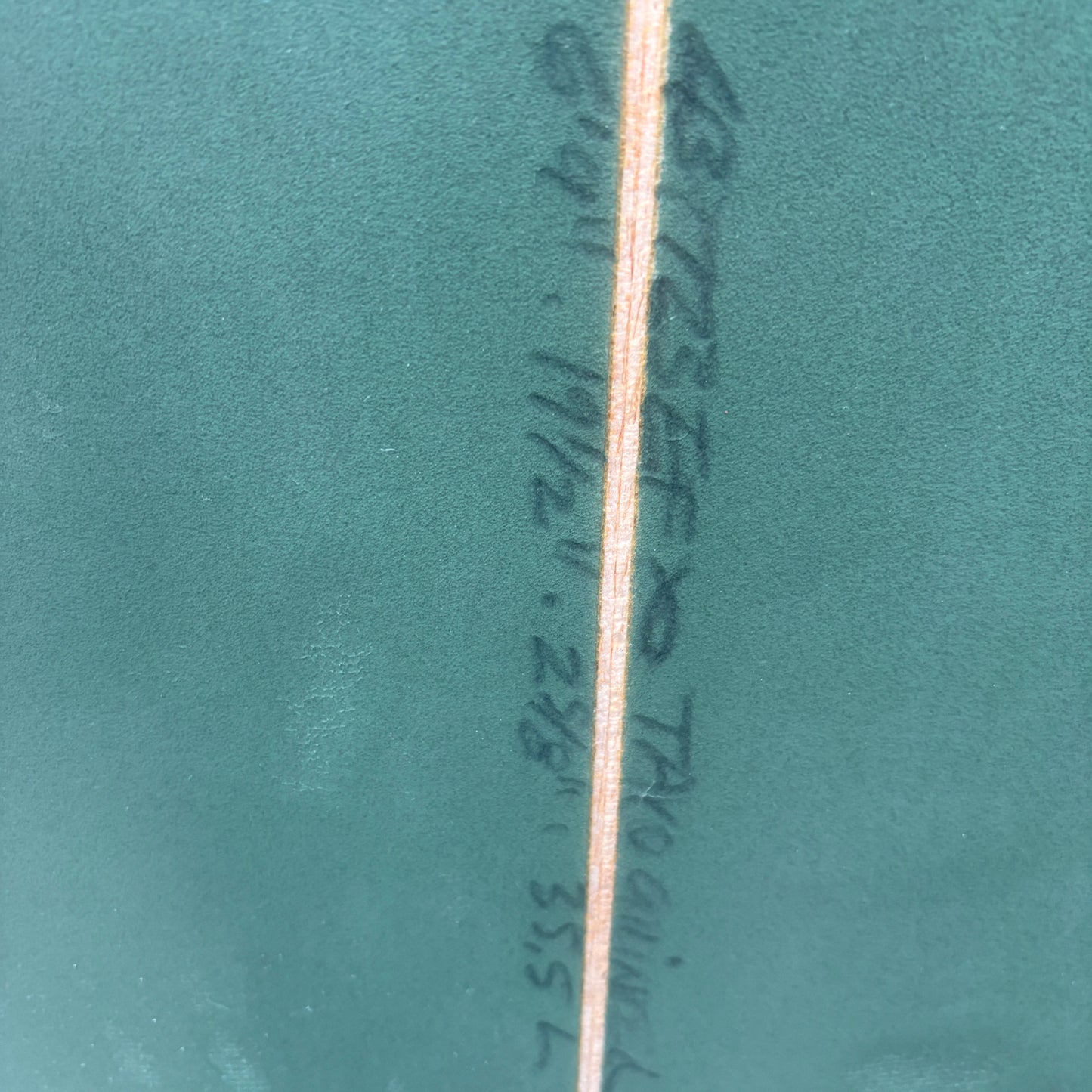 A close-up of a teal green textured surface with a vertical seam and handwritten black surfboard measurement markings in the center.