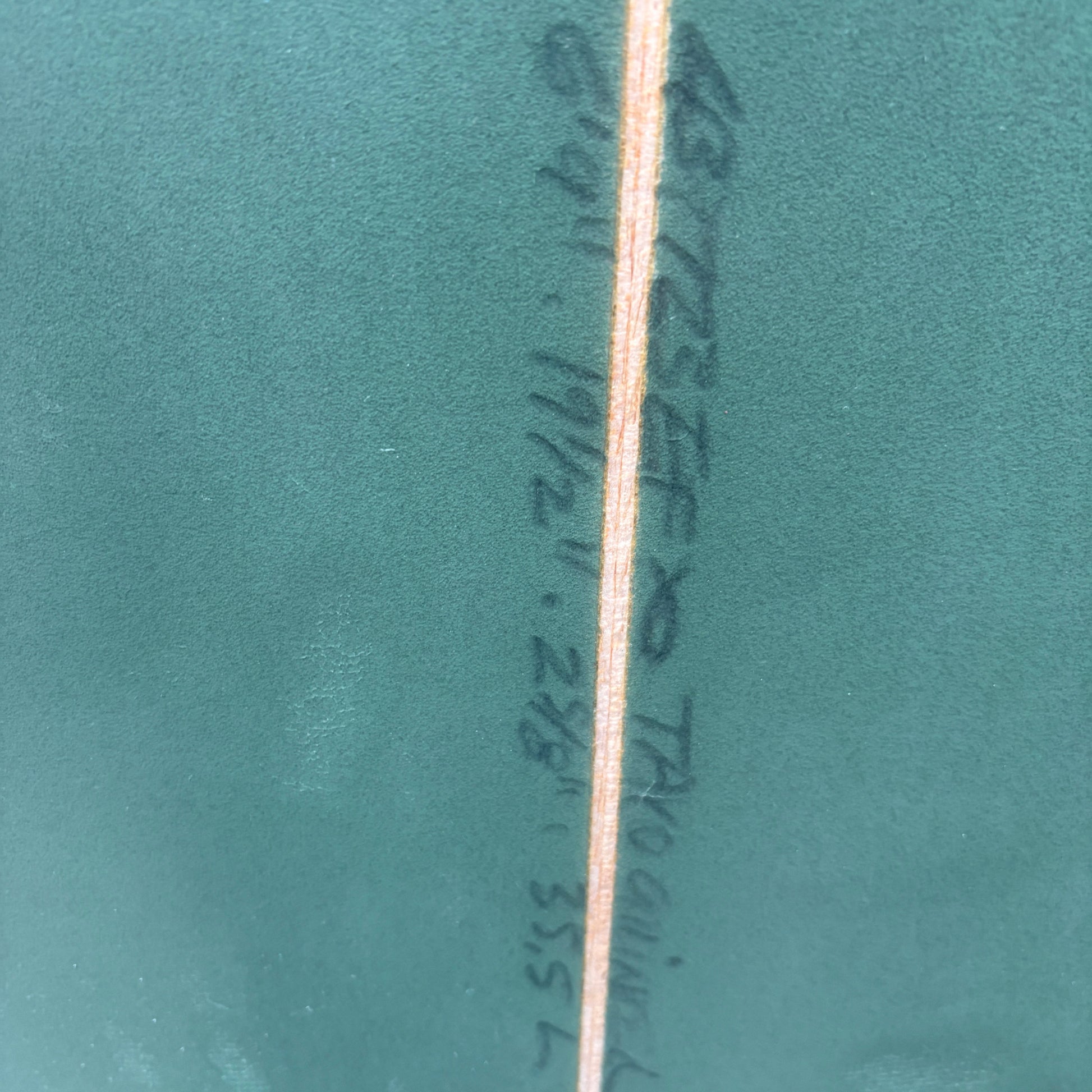 A close-up of a teal green textured surface with a vertical seam and handwritten black surfboard measurement markings in the center.