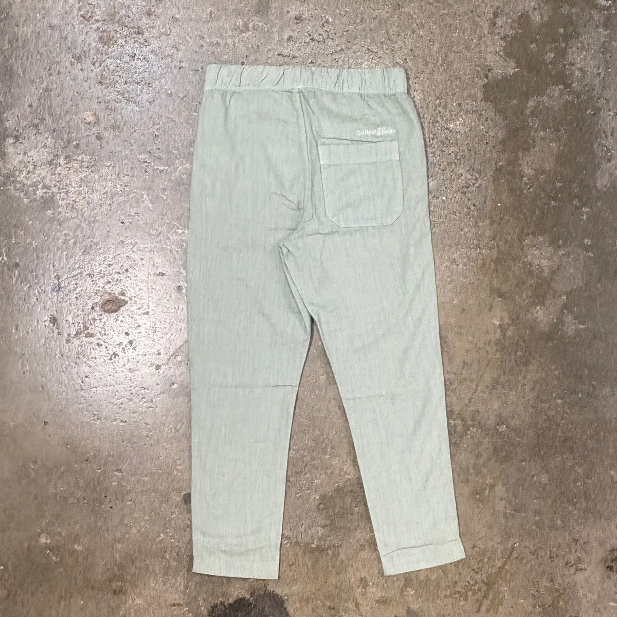 A pair of light sage green cropped linen pants with a back pocket, laid flat on a textured concrete floor.