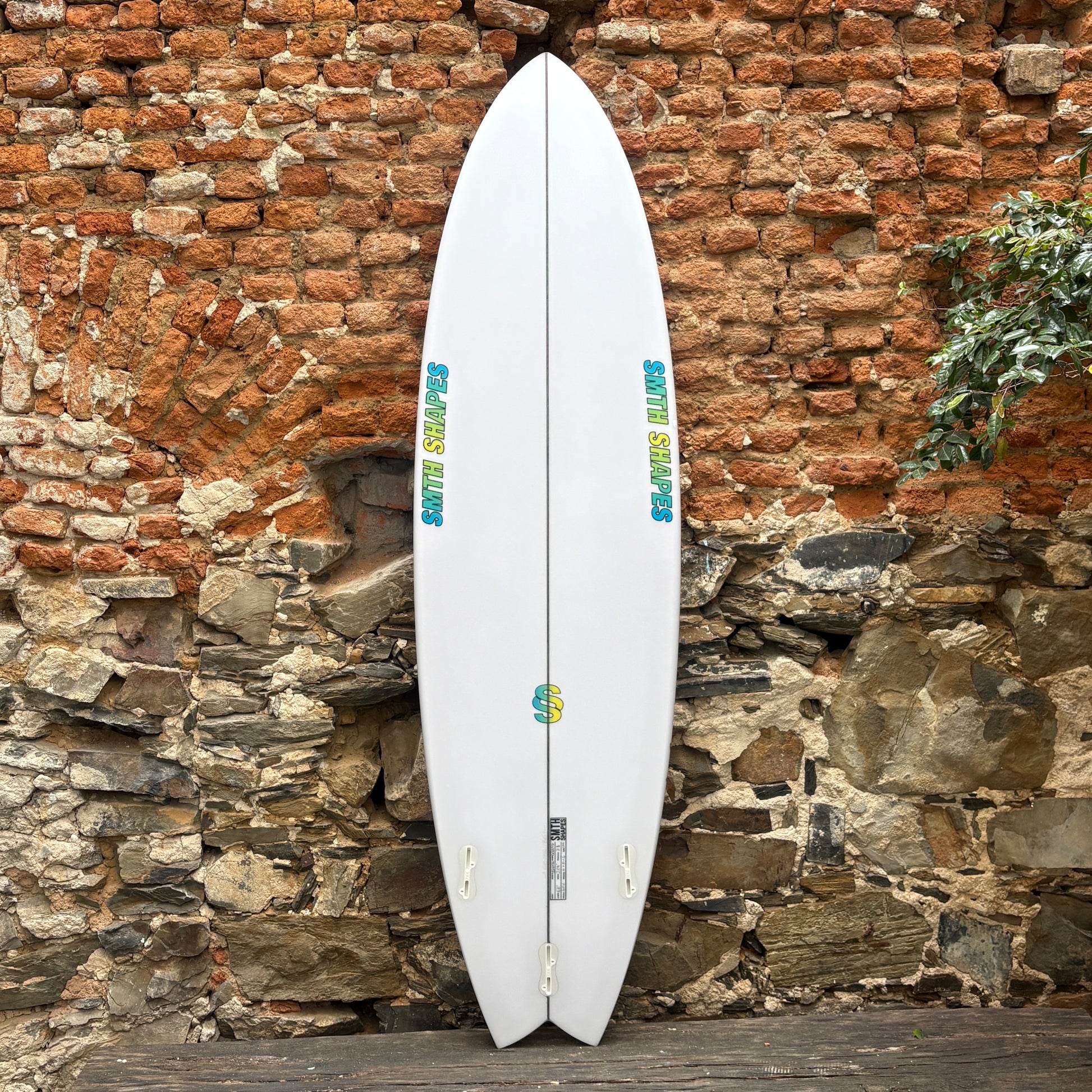 A white double-hulled surfboard with blue and yellow graphics, leaning against a rustic brick and stone wall.