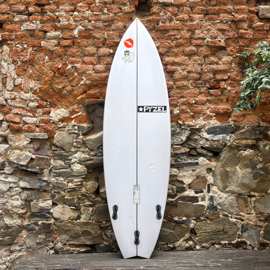 A white surfboard with black fins and stickers, including a prominent 'PYZEL' logo, leaning against a rustic brick and stone wall.