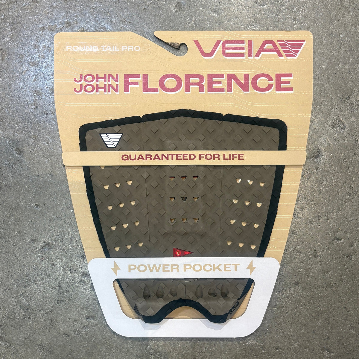 A Veia John John Florence surfboard grip pad in its original retail packaging, displayed on a textured gray concrete surface. The grip pad is olive brown with black trim and small beige triangles, and the backing card is beige with pink and white text.