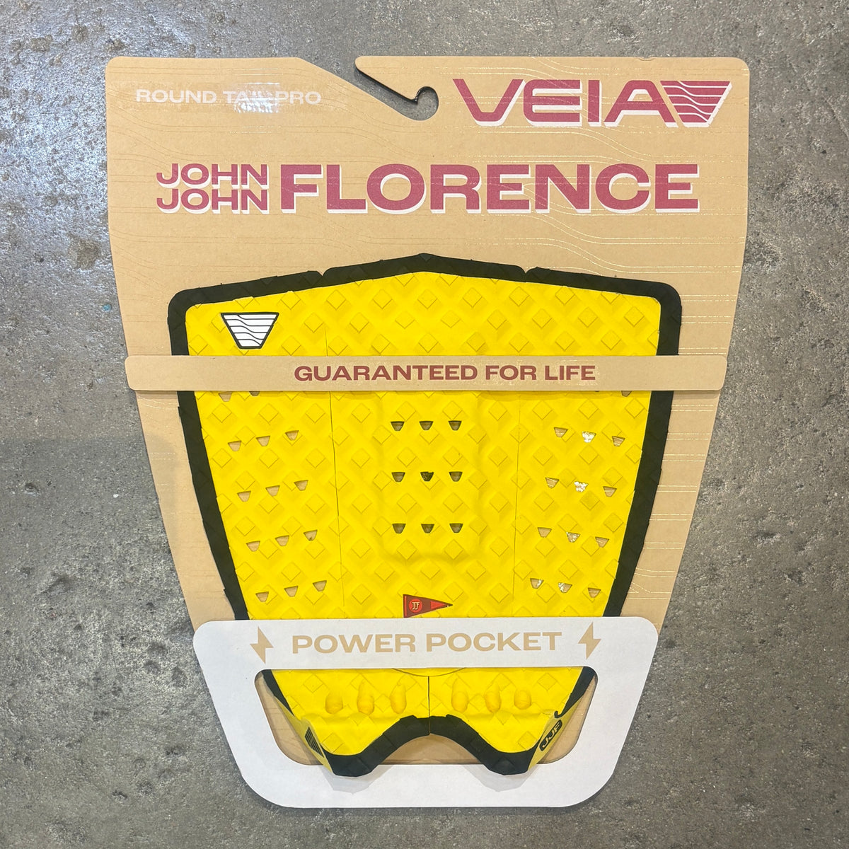 A yellow and black Veia John John Florence power pocket surfboard grip pad in its original cardboard packaging, displayed on a gray concrete surface.