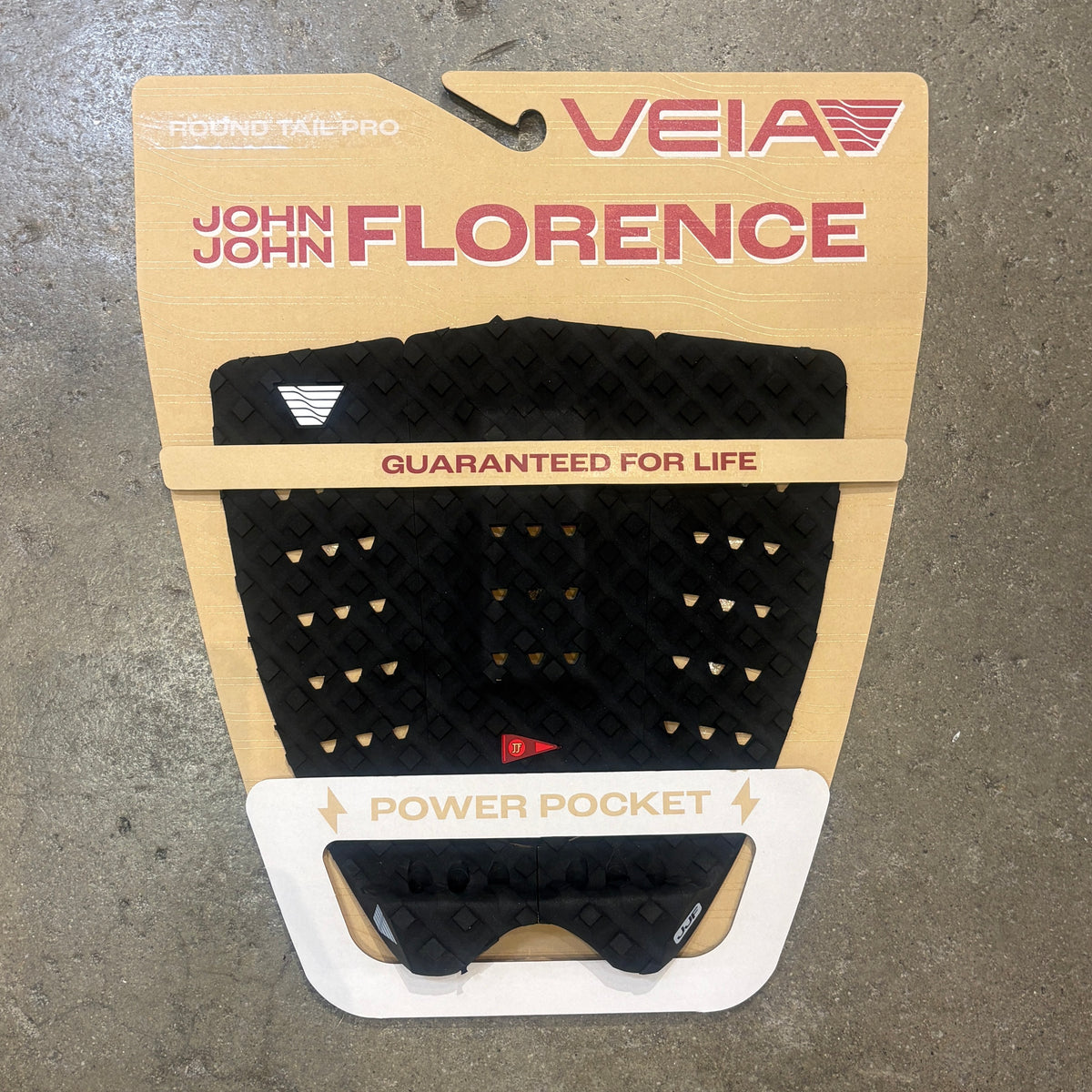 A black textured power pocket tail pad by Veia for John John Florence, mounted on its original tan cardboard retail card against a gray concrete floor.