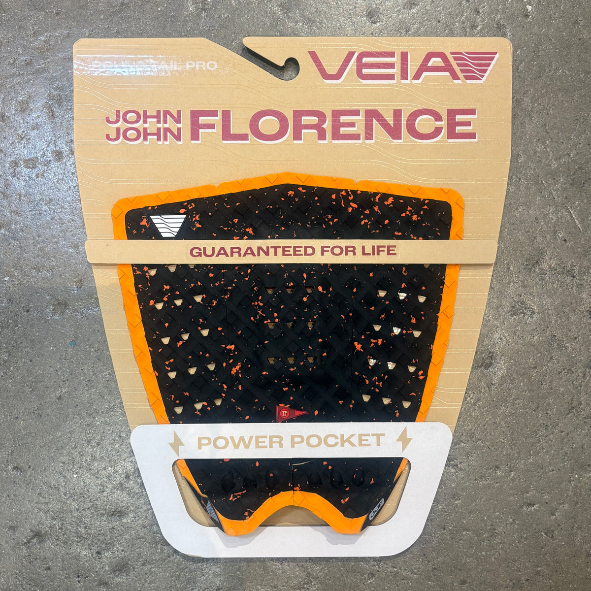 A Veia brand traction pad for a mountain bike, featuring a black textured surface with orange and white speckles, mounted on its original cardboard packaging over a gray concrete floor.