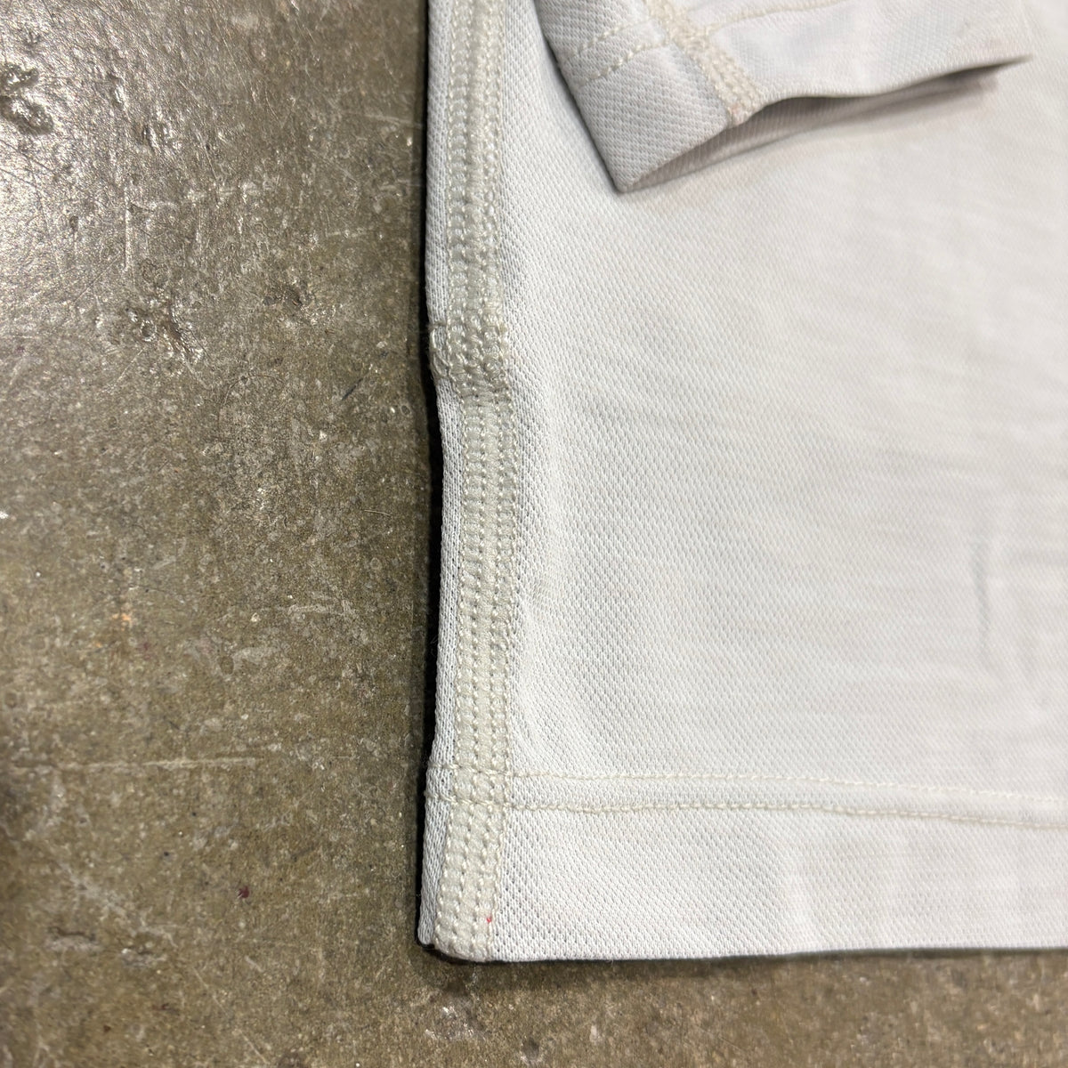 Close-up of a light off-white woven fabric corner with neat double stitching, laid flat on a textured gray concrete surface.