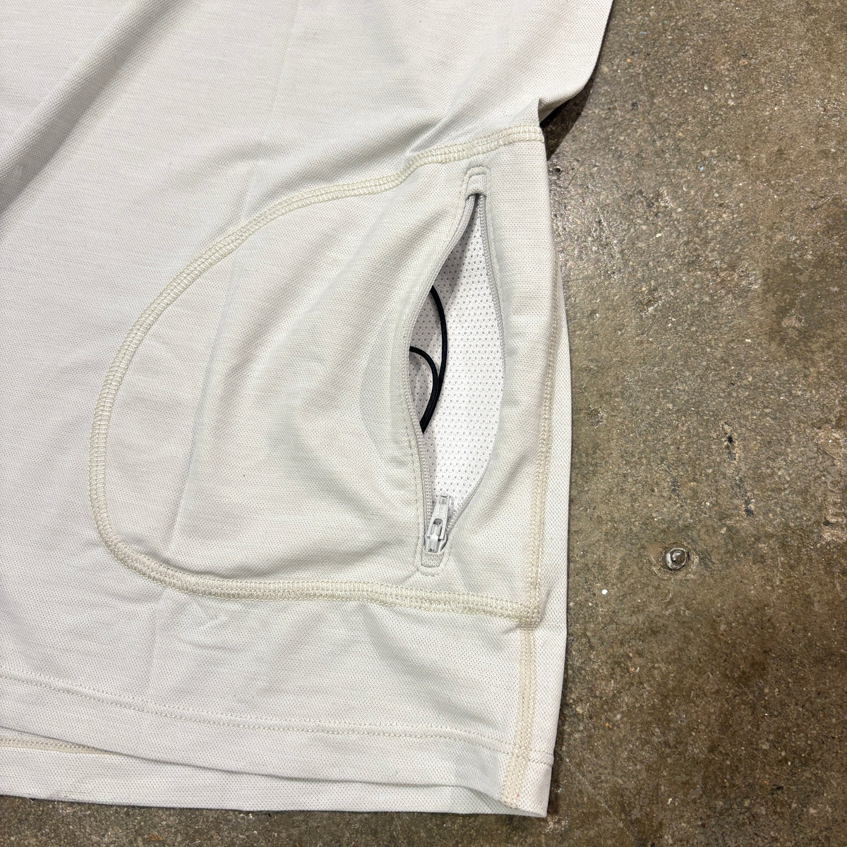 Close-up of a light beige or off-white athletic garment laid flat on a concrete floor, showing a zippered pocket opening with mesh lining.