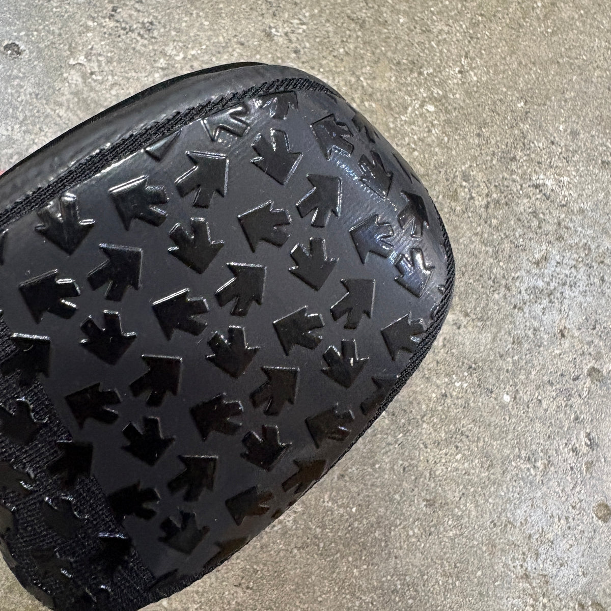 A close-up of a black textured bag or pouch with a repeating embossed arrow pattern, photographed on a concrete floor.