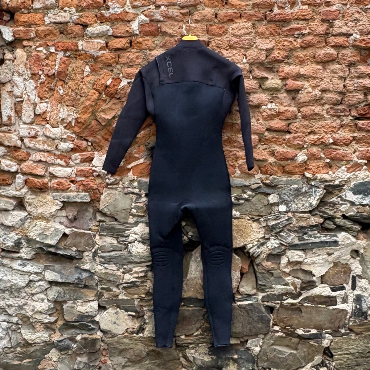 A black full-body wetsuit hanging on a yellow hanger against a rustic brick and stone wall.