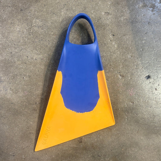 A single swim fin with a two-tone color scheme of royal blue and bright yellow, photographed on a textured gray concrete floor. The brand name 'GALERA' is embossed near the bottom tip.