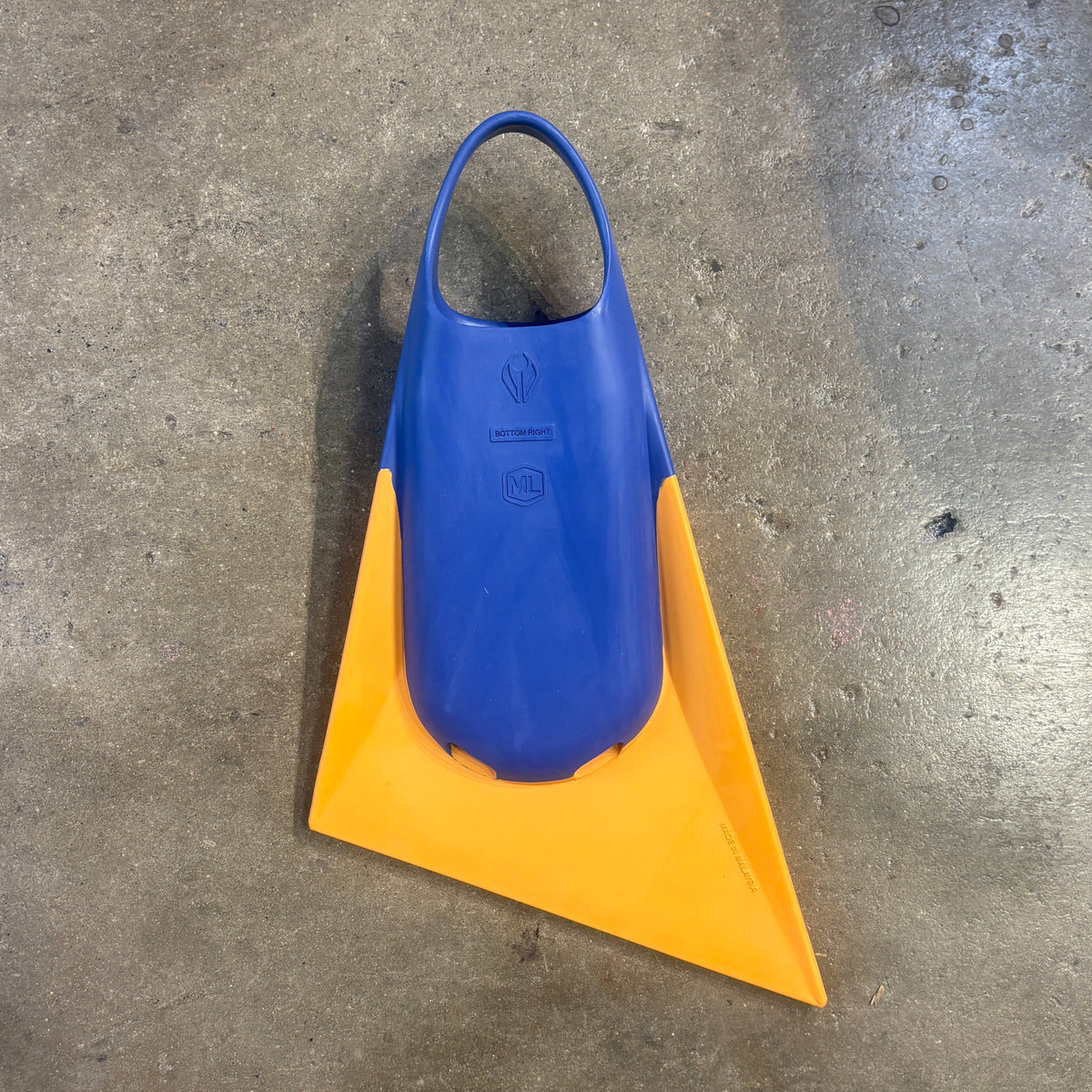 A bottom view of a single swim fin with a blue upper and a bright yellow lower blade, photographed from above on a gray concrete floor.