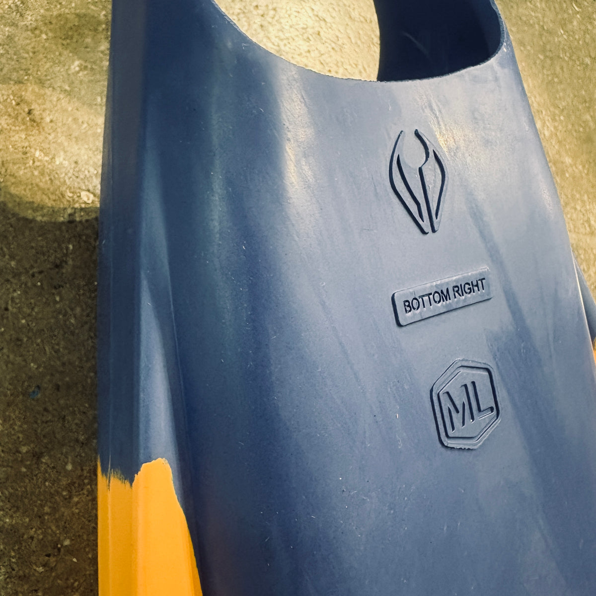 Close-up of a dark blue molded plastic component with yellow accents, featuring embossed logos and a label reading 'BOTTOM RIGHT' on an outdoor concrete surface.