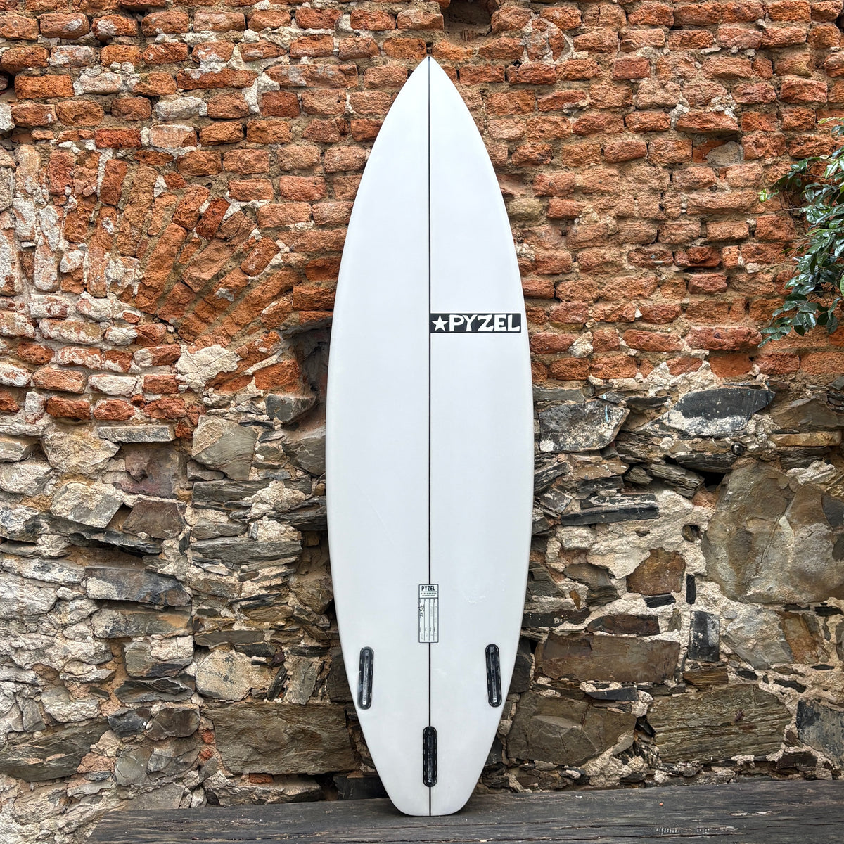 A white surfboard with a black PYZEL logo, leaning vertically against a rustic brick and stone wall.