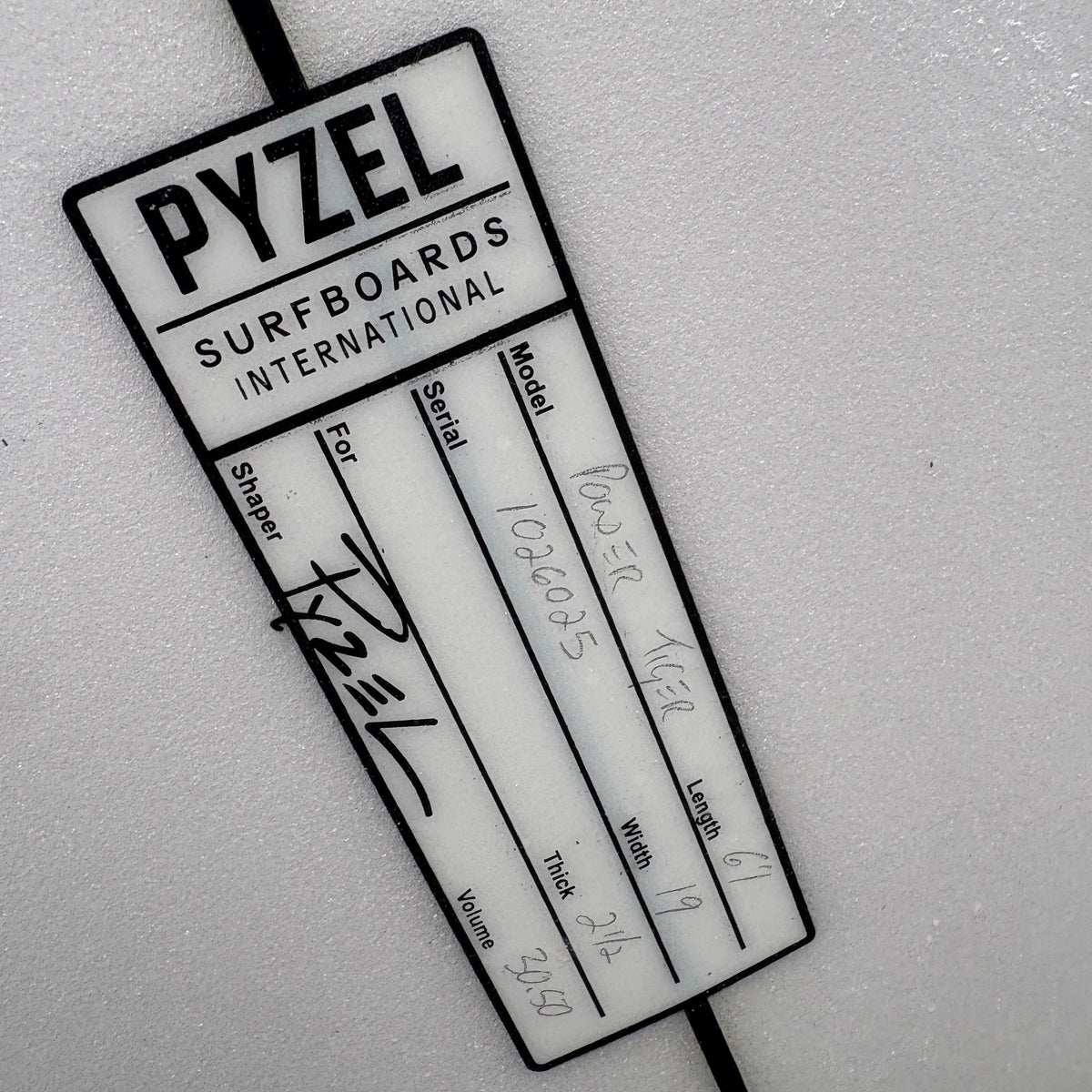 A white and black Pyzel Surfboards International identification tag attached to a black strap, photographed on a light textured surface.