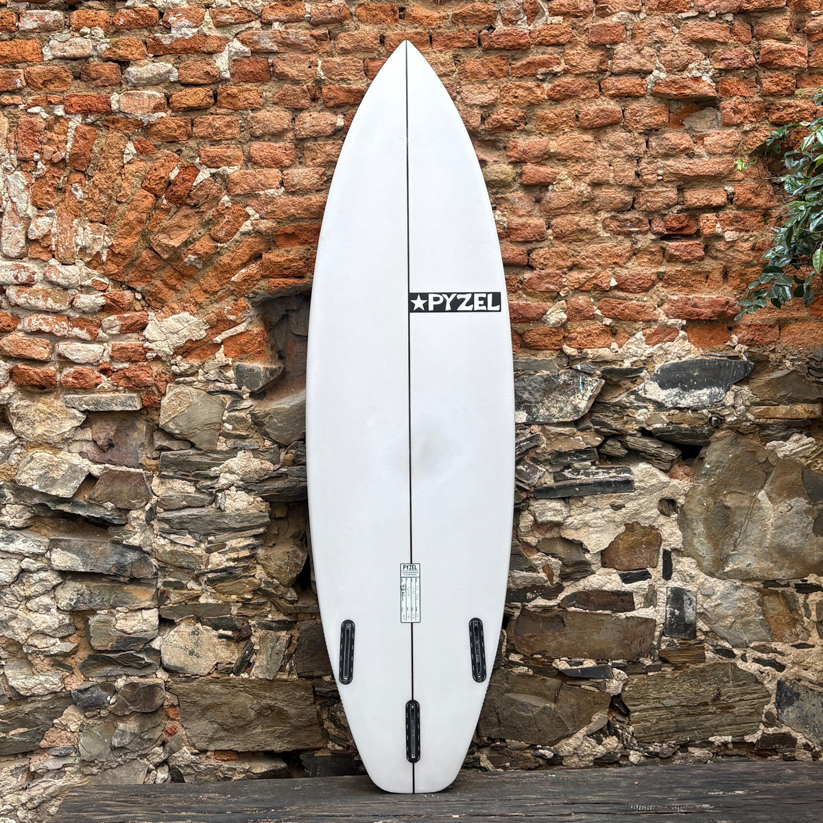 A white Pyzel surfboard standing upright against a rustic brick and stone wall.