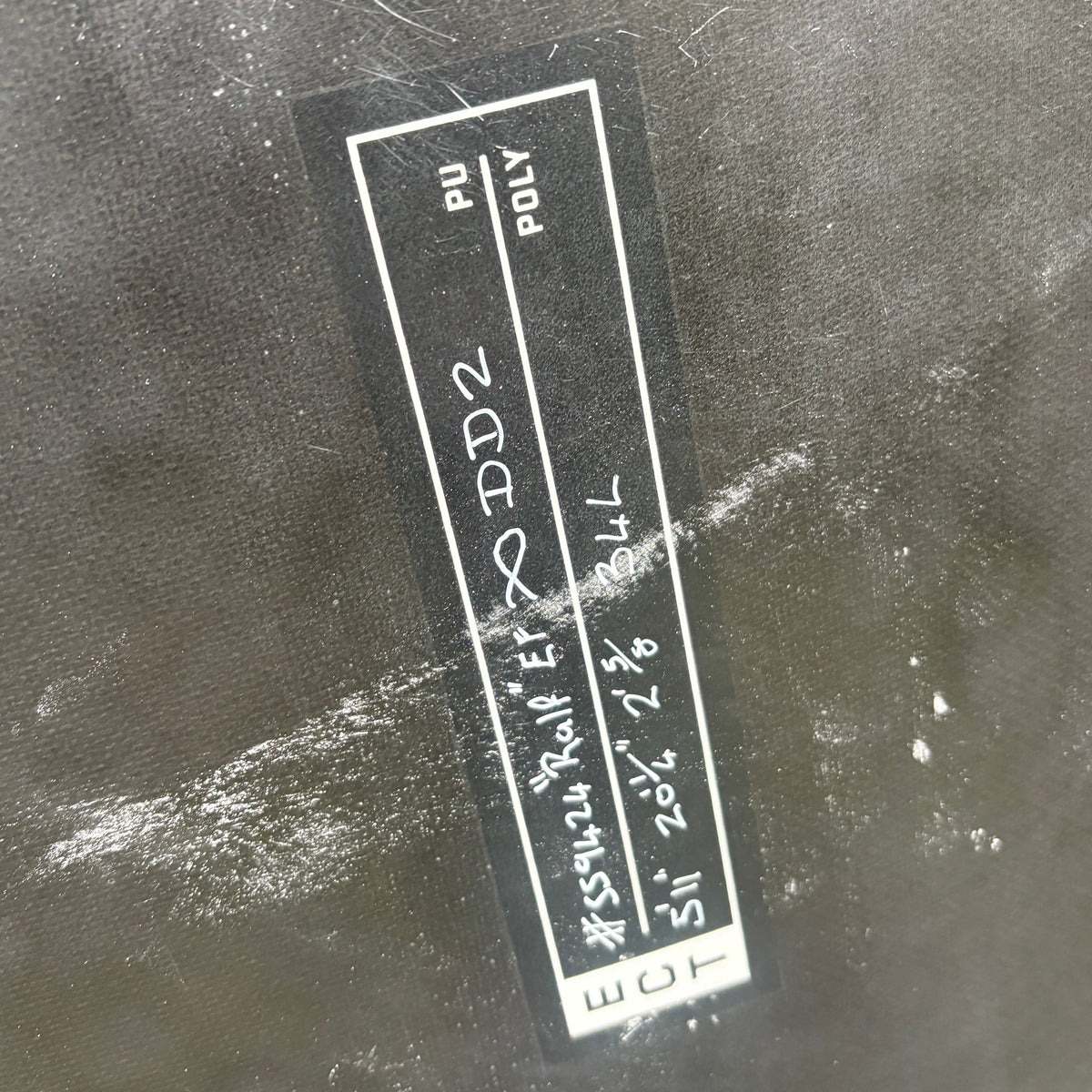 A close-up of a black fabric item with a sewn-on black label that reads 'DD2 POLY PIU' and includes handwritten size information in white text.