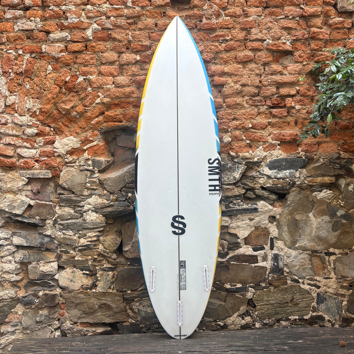 A white Smith surfboard with blue and yellow accents, standing vertically against a rustic brick and stone wall.