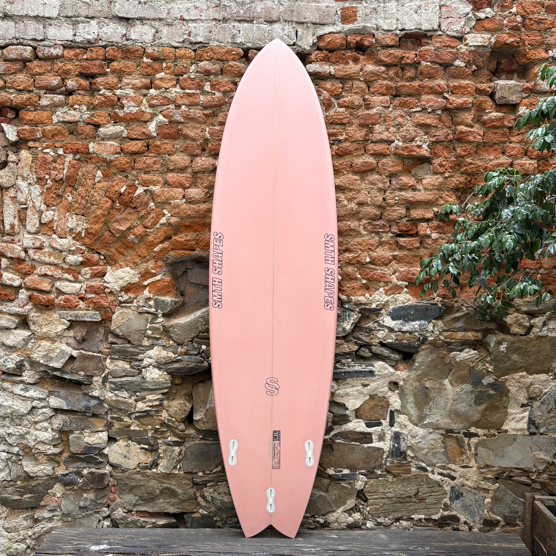 Pink surfboard leaning against a rustic stone wall with a brick background.