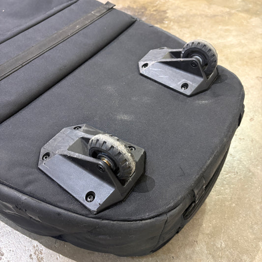 Close-up of the bottom corner of a black soft-sided bag or case, showing two black caster wheels mounted on plastic bases on a concrete floor.