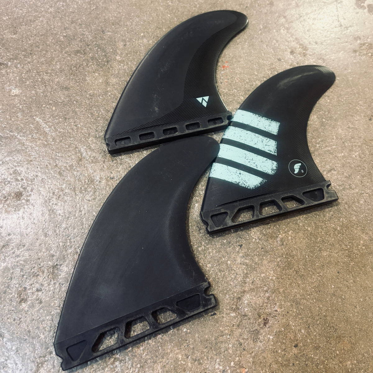 Three black carbon fiber surfboard fins with white graphics, including stripes and a small logo, displayed on a concrete floor.