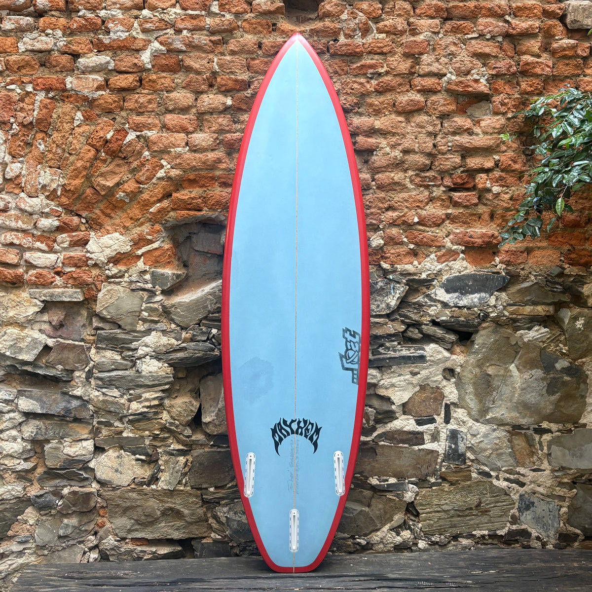 A light blue surfboard with a red border and black graphics, standing upright against a rustic brick and stone wall.