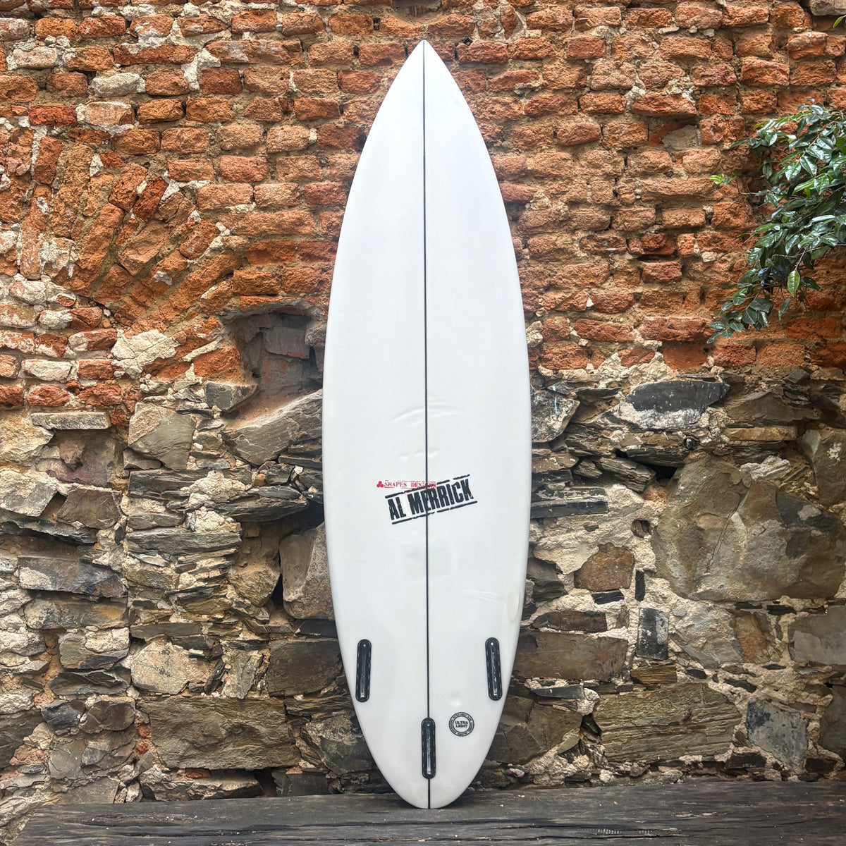 A white surfboard with black fins and a red and black Al Minnick logo, leaning against a rustic brick and stone wall.