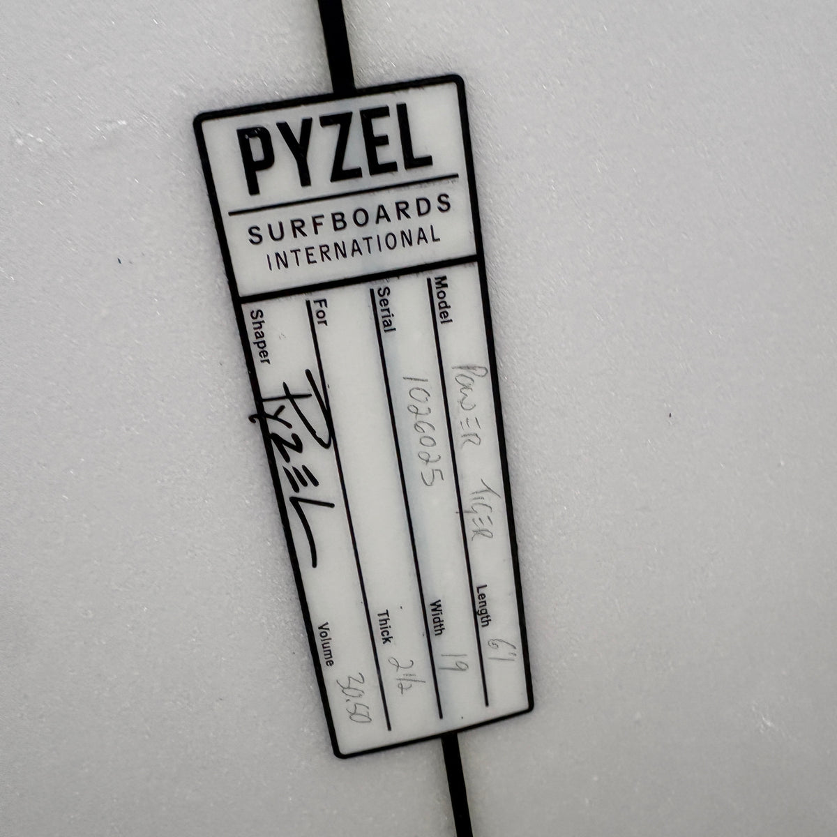 A white Pyzel Surfboards International identification card or label with black printed text and handwritten markings, held by a black string against a light gray background.