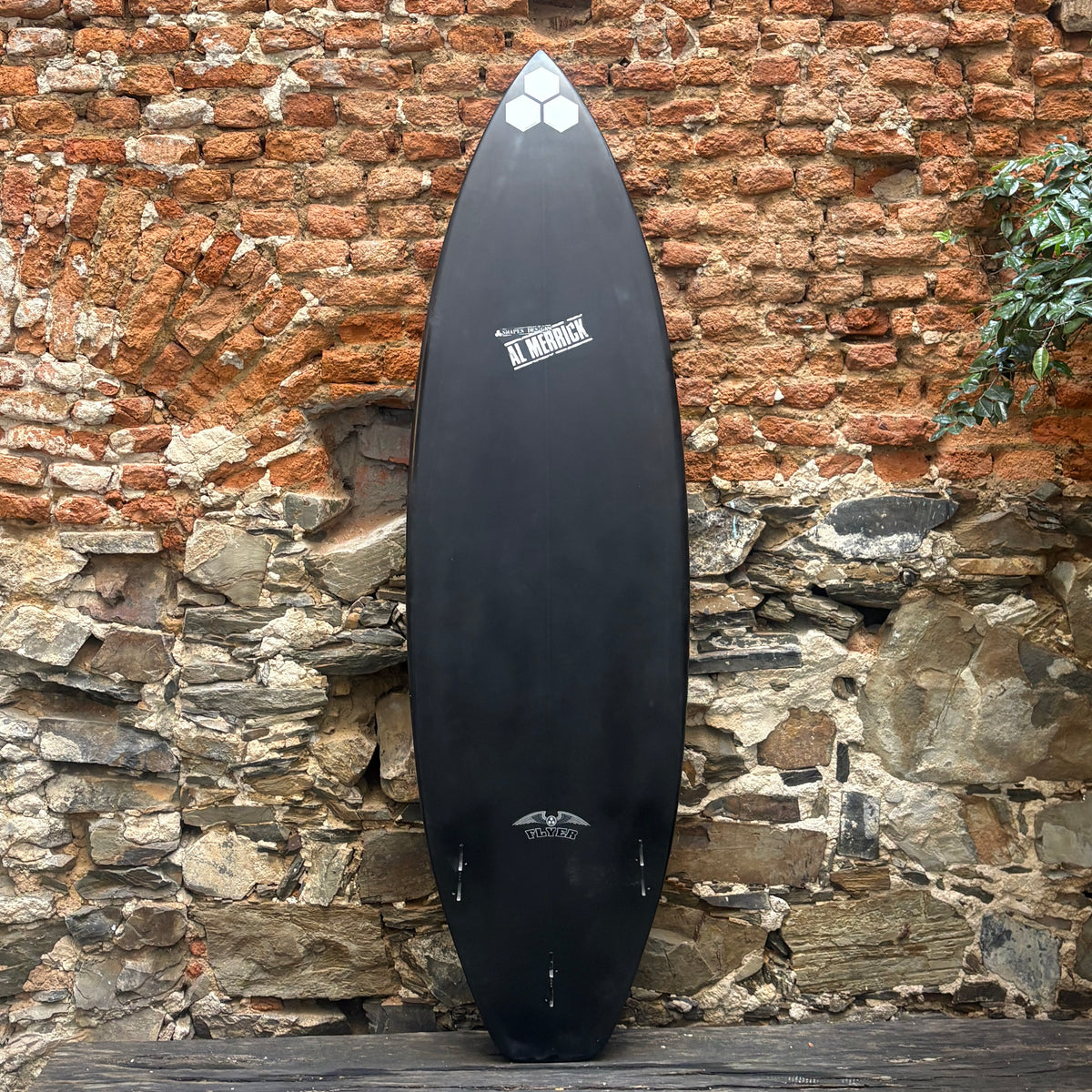 A black surfboard with white graphics, leaning against a rustic brick and stone wall.