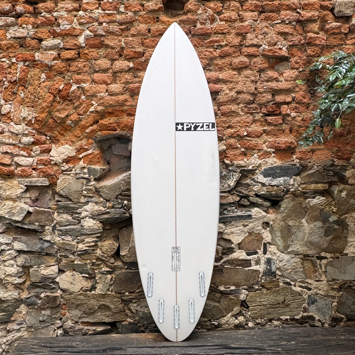 A white Pyzel surfboard standing upright against a rustic brick and stone wall.