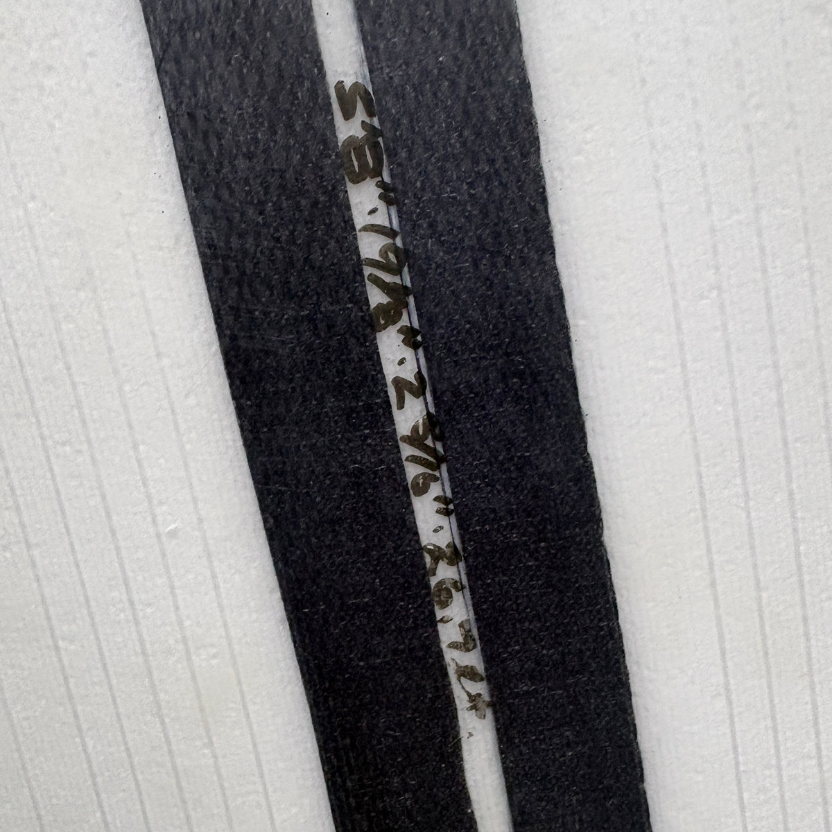 A close-up of a black fabric strap or trim laid diagonally across a white textured background, with handwritten measurement markings in black ink along the center.