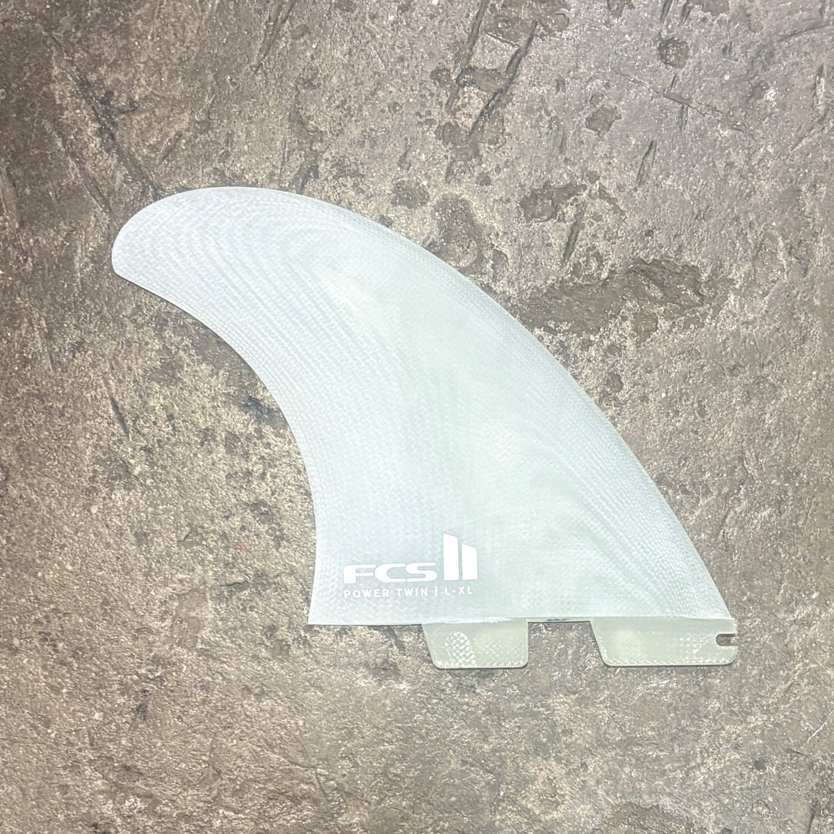 A translucent white surfboard fin blank with a visible FCS logo, photographed flat on a textured gray stone surface.