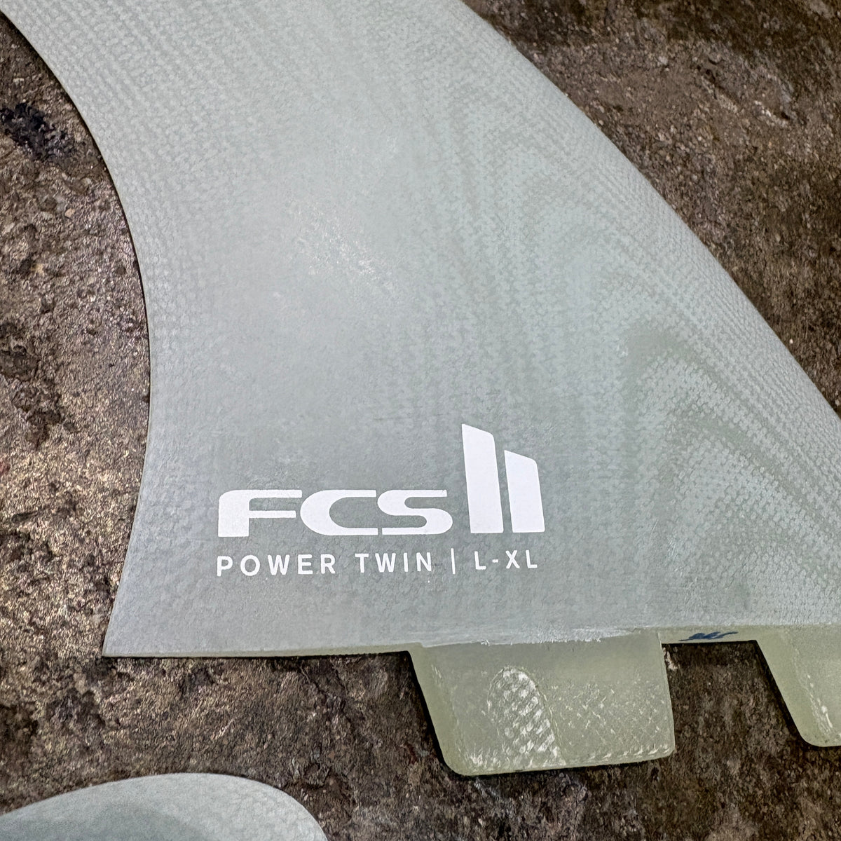 A close-up of a translucent gray surfboard fin with a white printed FCS logo and the text 'POWER TWIN | L-XL' on a textured stone surface.