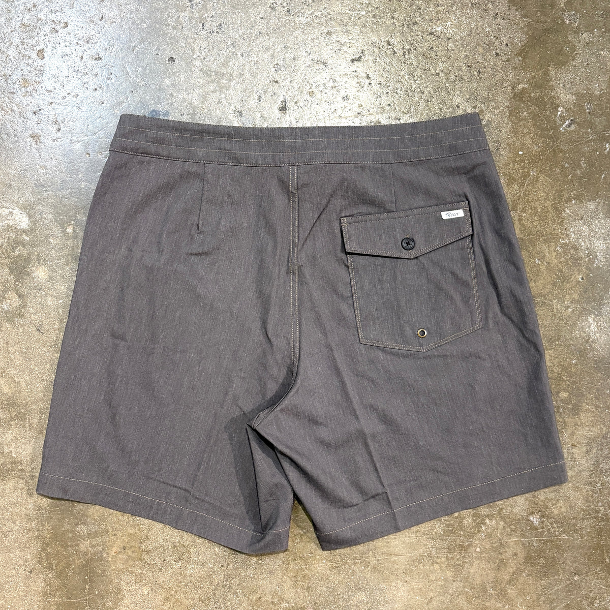 A pair of dark gray chambray shorts laid flat on a textured concrete floor, showing the back pocket with a button flap.